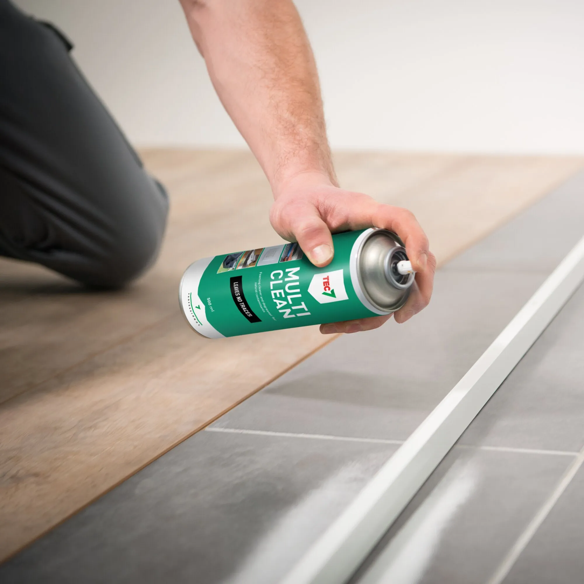 Hand hält eine grüne Sprühdose Schaumreiniger MULTICLEAN über einem Boden aus Holz- und grauen Fliesenflächen