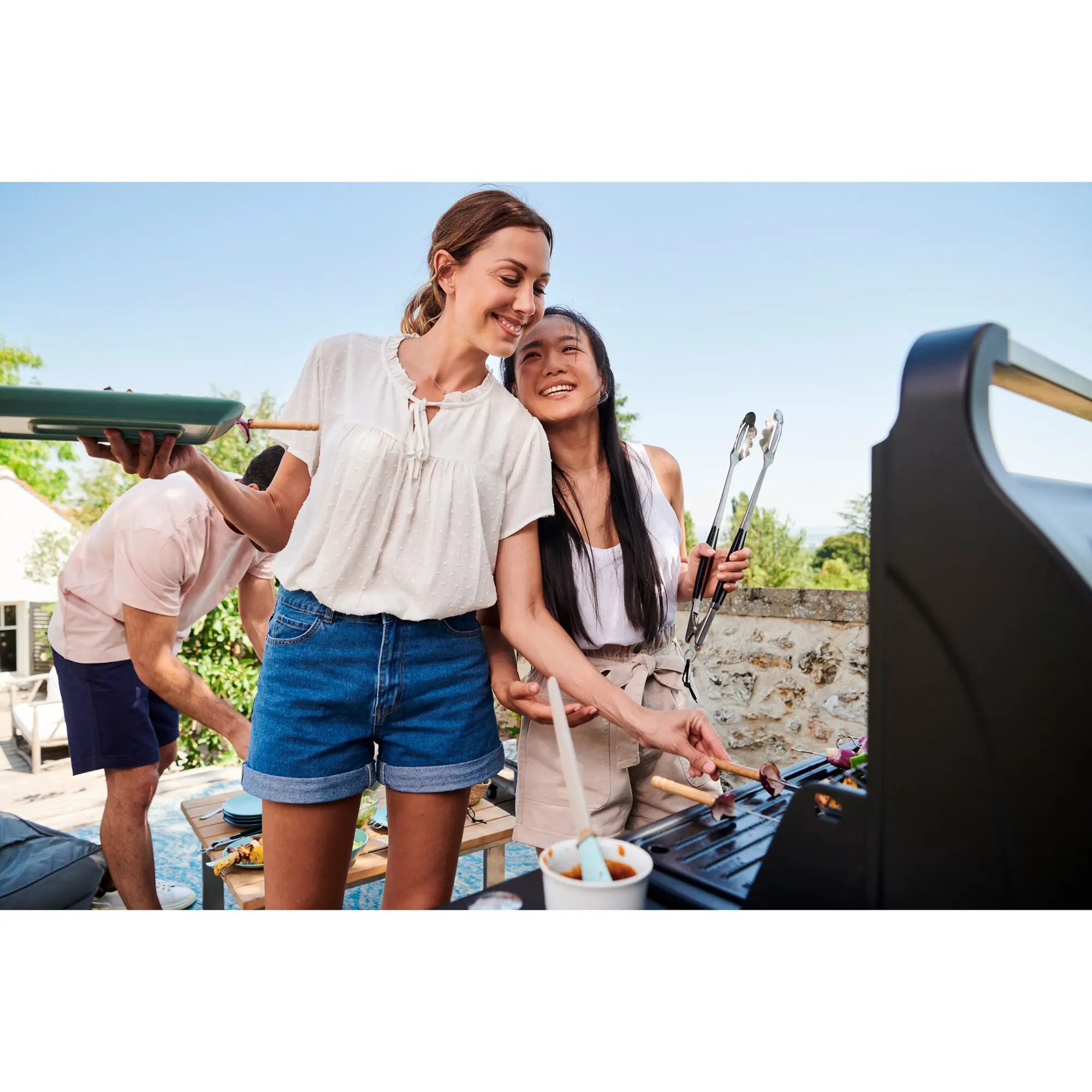 Fröhliche Gruppe beim Grillen im Freien; rechts der Gasgrill Compact 3 L mit Haube, zwei Frauen halten Spieße und genießen Essen