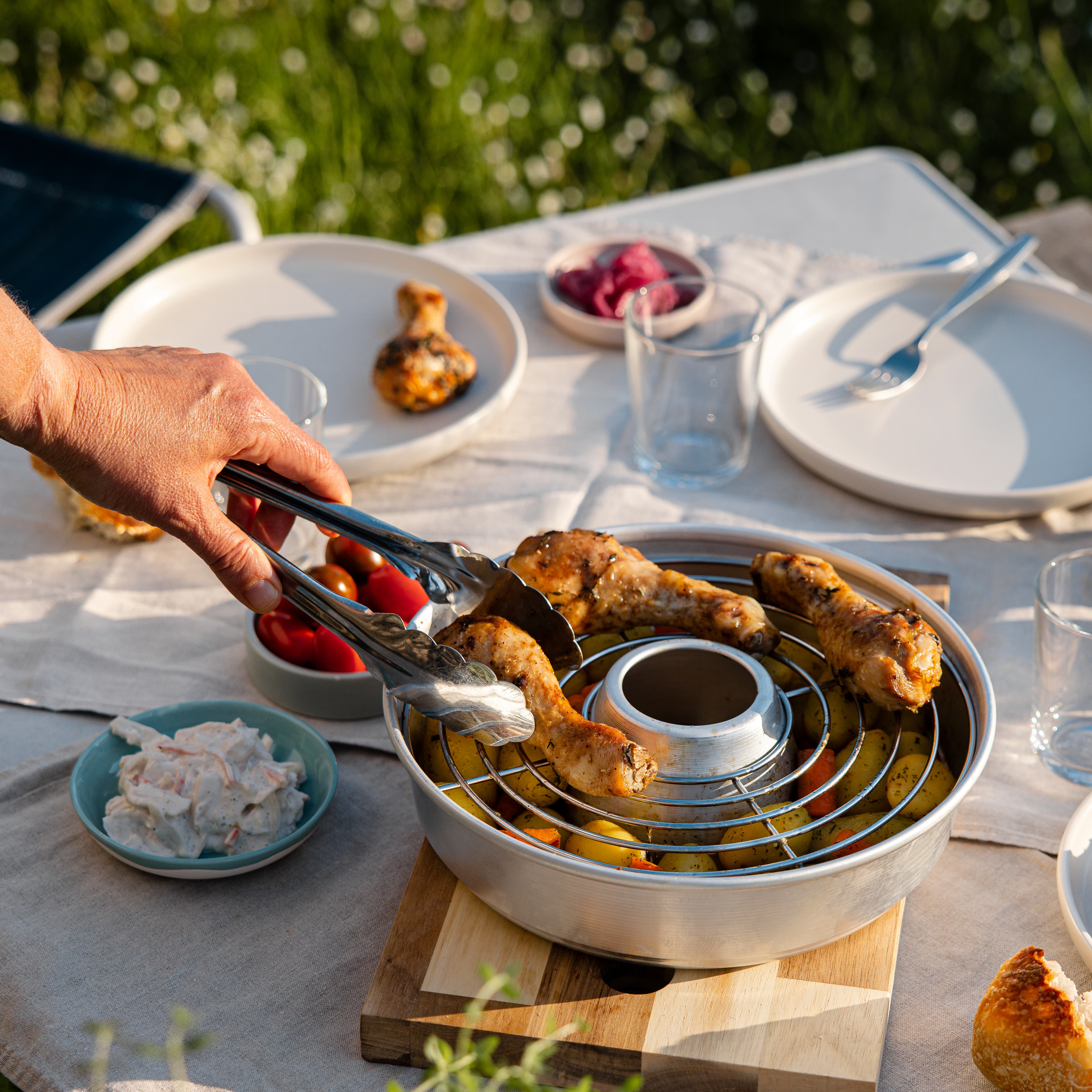 Aufbackgitter Ø 22,5 cm als runder Metallkorb mit Ringen auf Holzunterlage; eine Hand greift gegrillte Hähnchenteile, daneben Tomaten und Kartoffeln