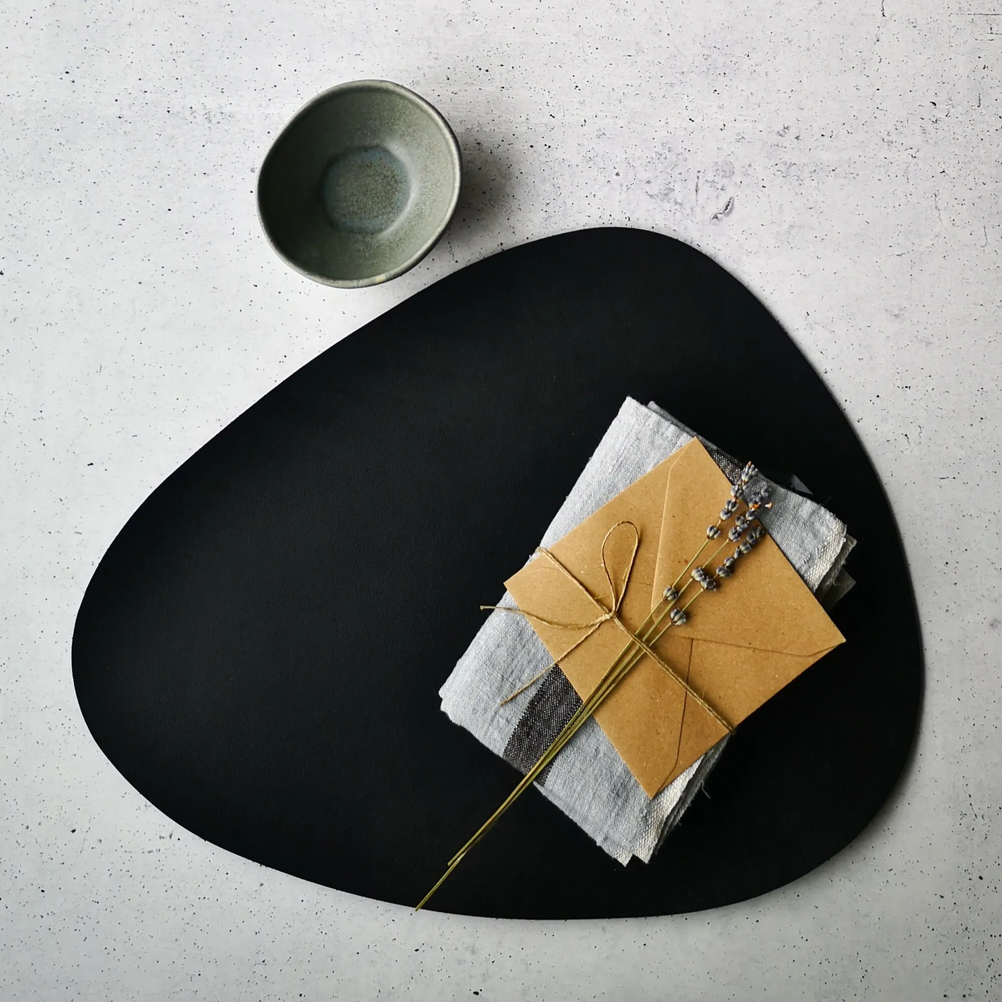 Schwarzes ovales Platzset auf hellem Tisch, dekoriert mit Serviette, Kraftpapier, Lavendelzweigen und kleiner Schale