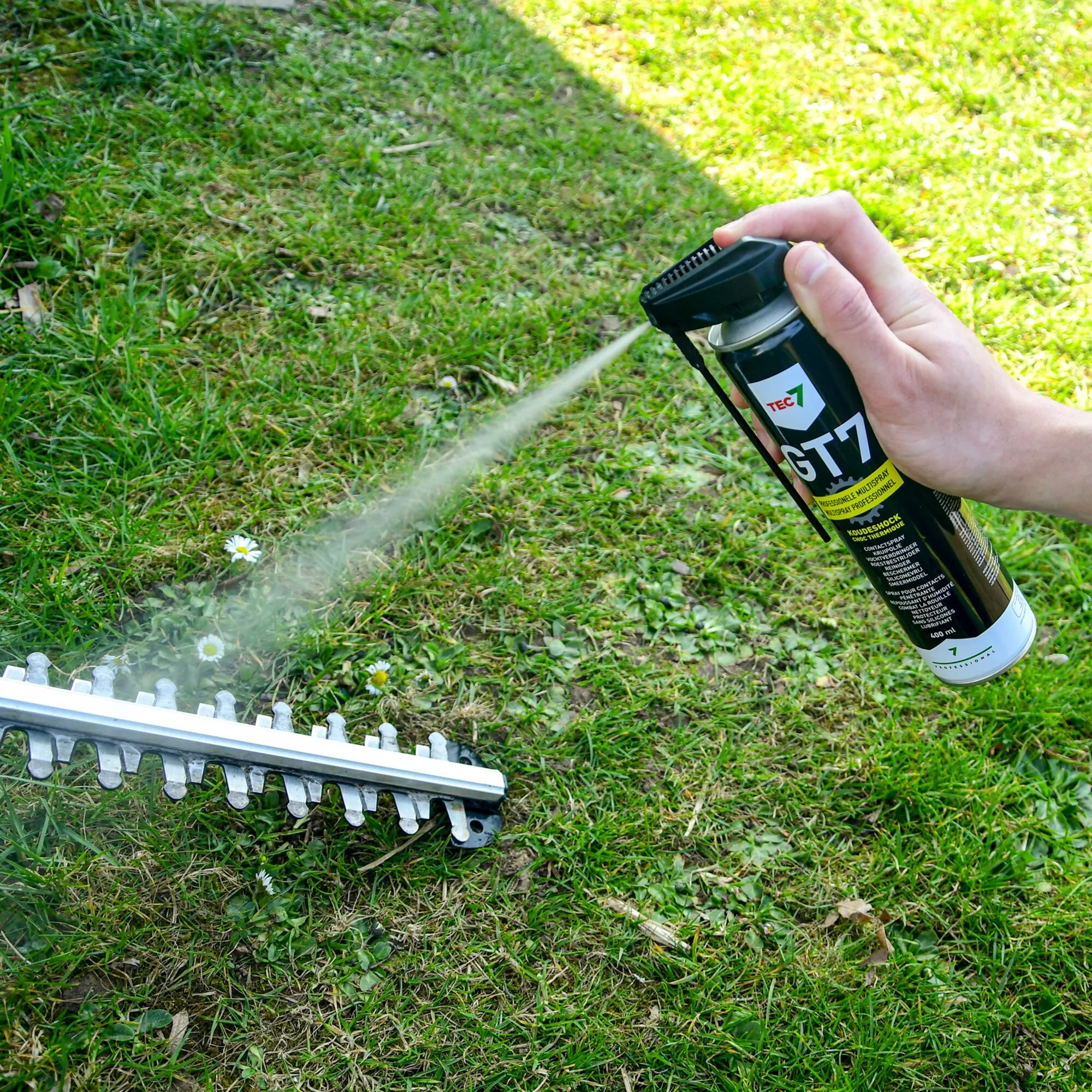 Hand hält schwarzes GT7 Multifunktionsspray und sprüht es auf eine auf dem Rasen liegende Heckenscherecuntegn