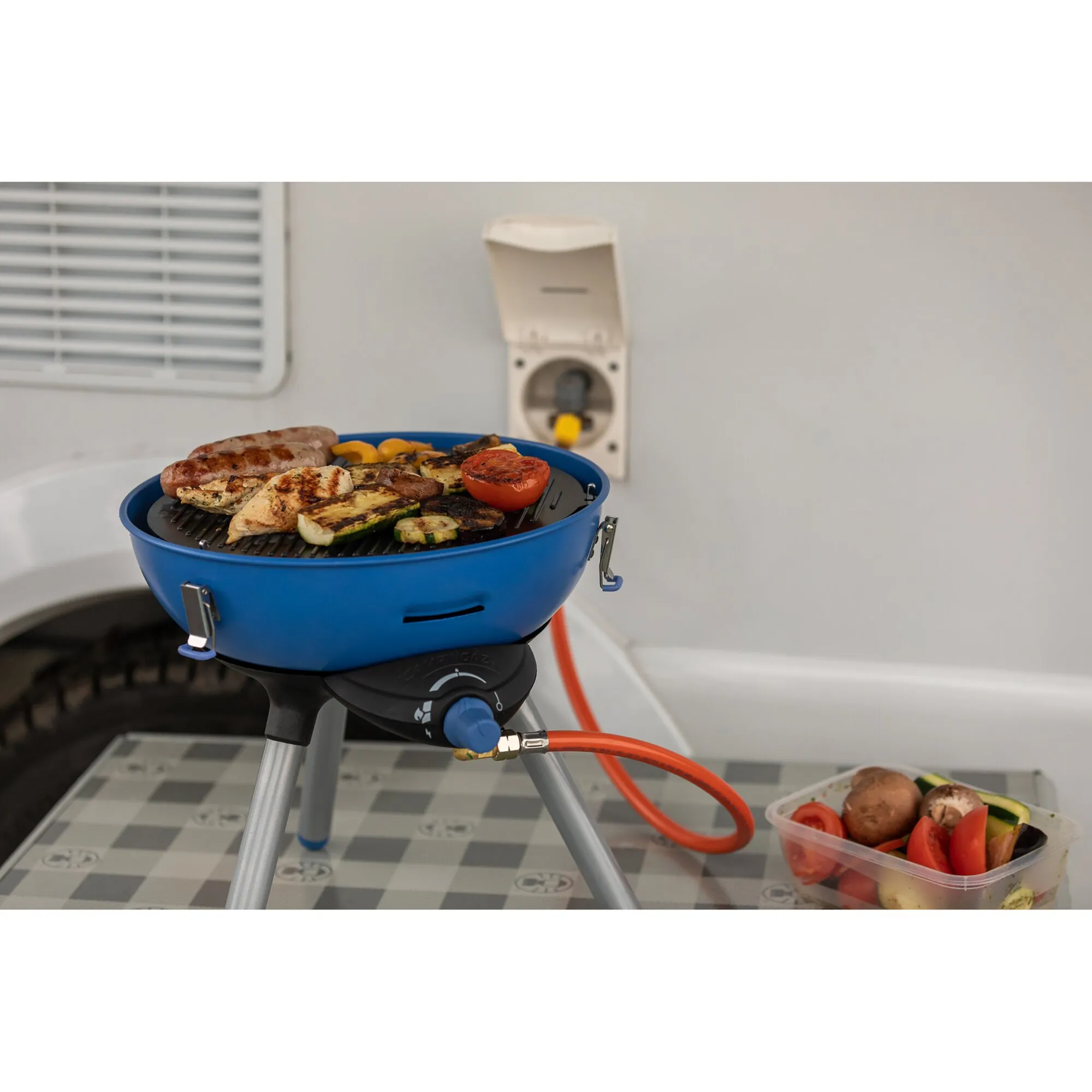 Blauer Party Grill 400 Caravan Connect auf Standbeinen mit gegrilltem Fleisch und Gemüse, angeschlossen an eine Gasflasche