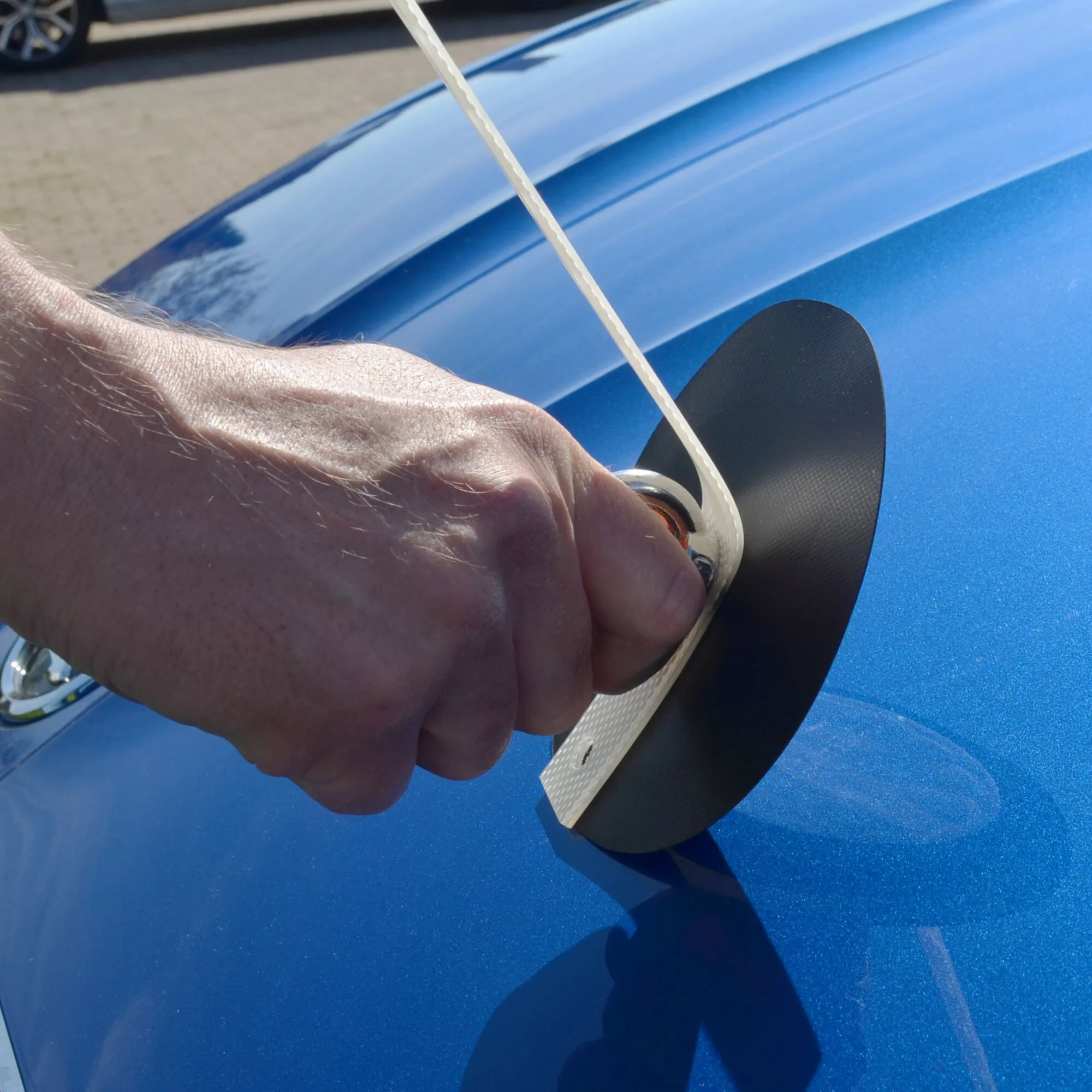Detailaufnahme der flexiMAGS-Magnetbefestigung für ein Sonnensegel: Eine Hand fixiert weißen Gurt am blauen Autodach