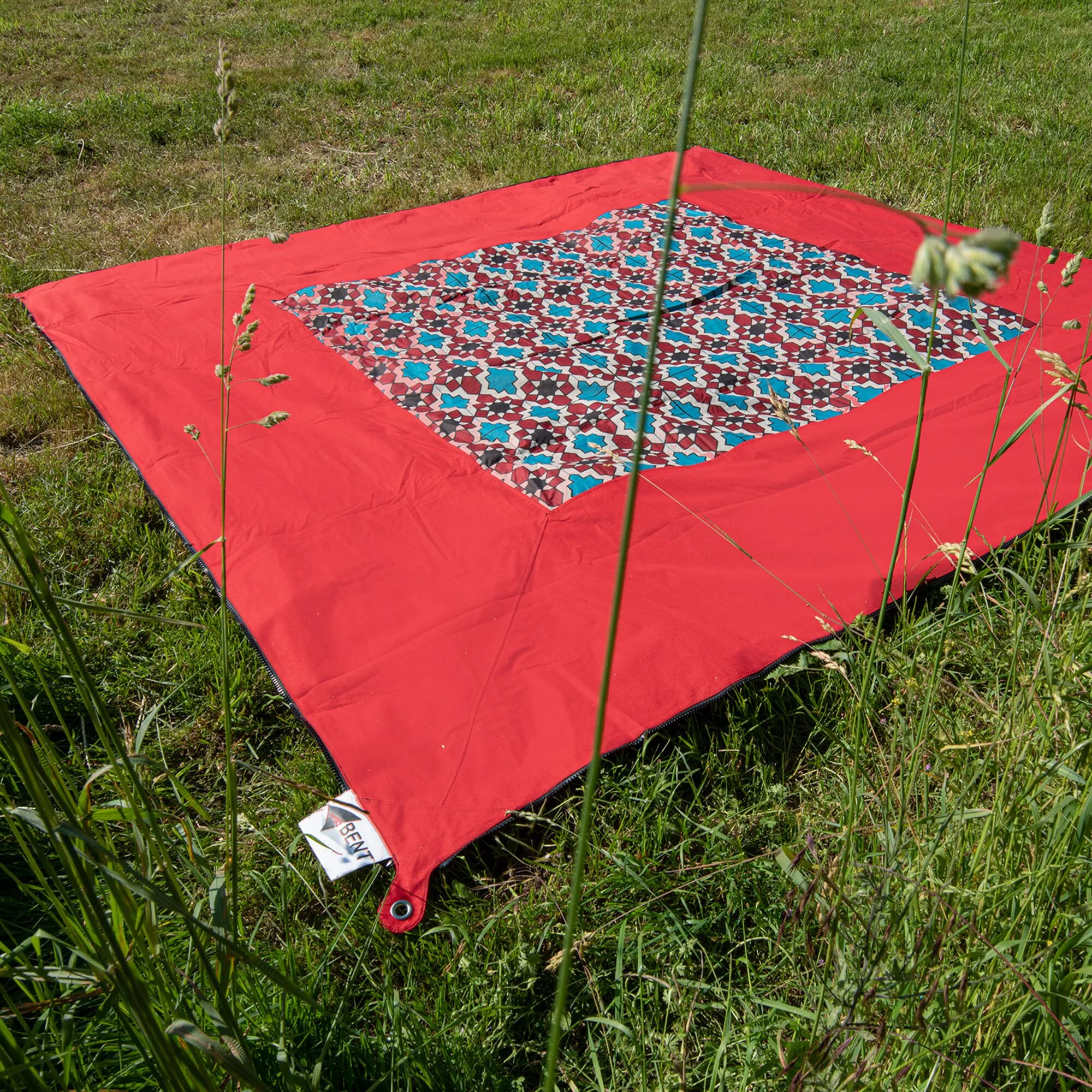 Roter Zip Carpet auf einer Wiese mit farbenfrohem geometrischem Muster in der Mitte und Öse an der Ecke