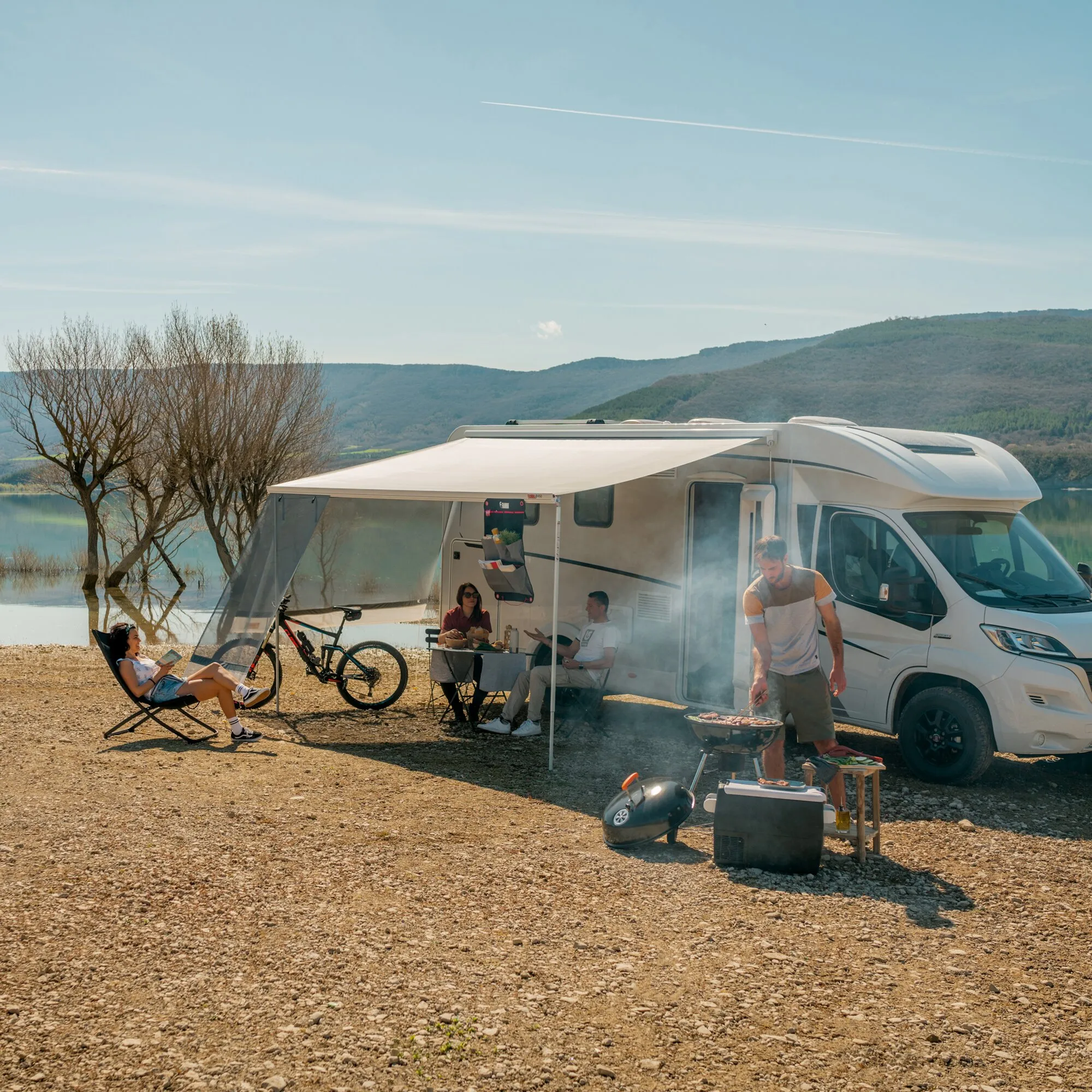Wohnmobil mit ausgefahrener Seitenwand-Markise Sun View Side; draußen sitzen Personen am Tisch, daneben Fahrräder und ein Grill, Hügel im Hintergrund
