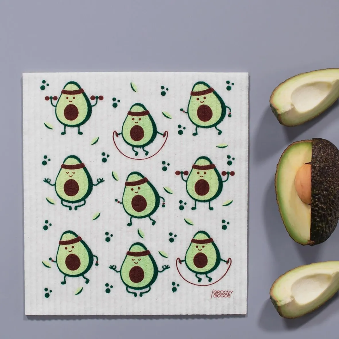 Ökologisches Schwammtuch mit Avocado-Muster und tanzenden Avocadofiguren, daneben halbierte Avocados auf grauem Hintergrund