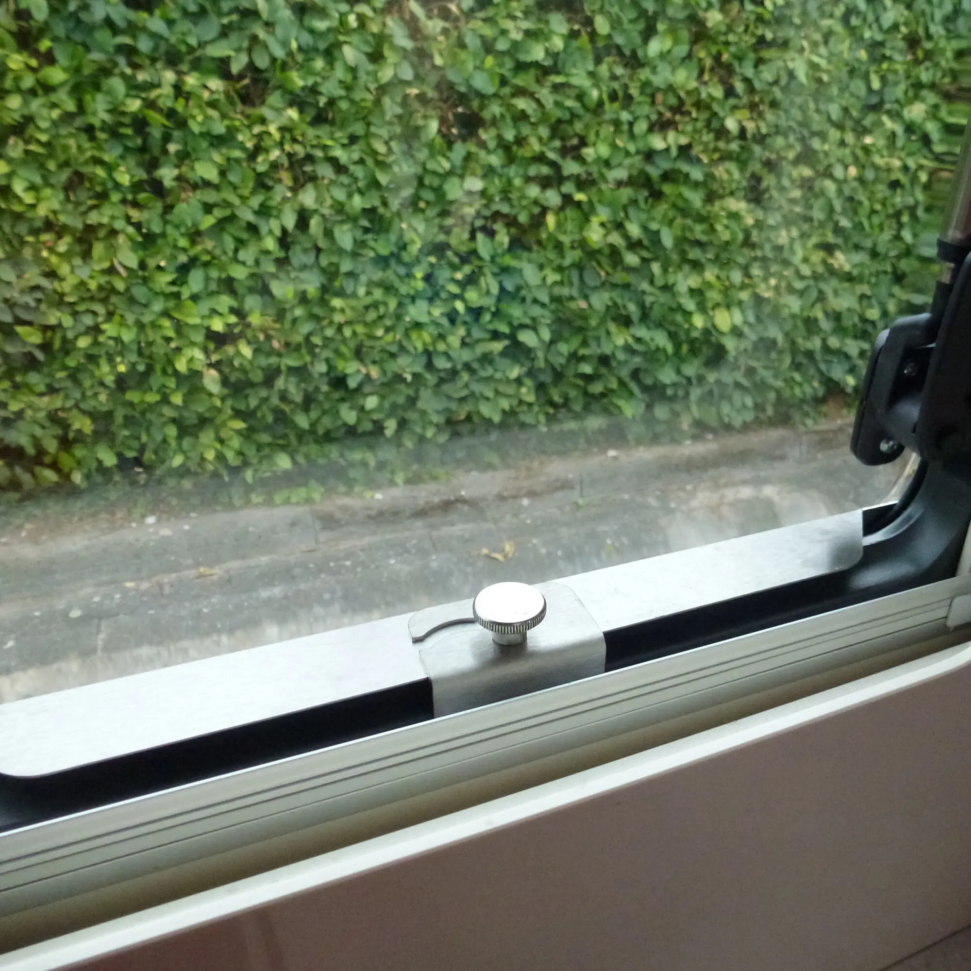 Innenansicht eines Fensters mit Sicherheitsprofil am Fensterrahmen, runder Griffknopf und gebogene Halterung, Blick auf eine grüne Hecke draußen