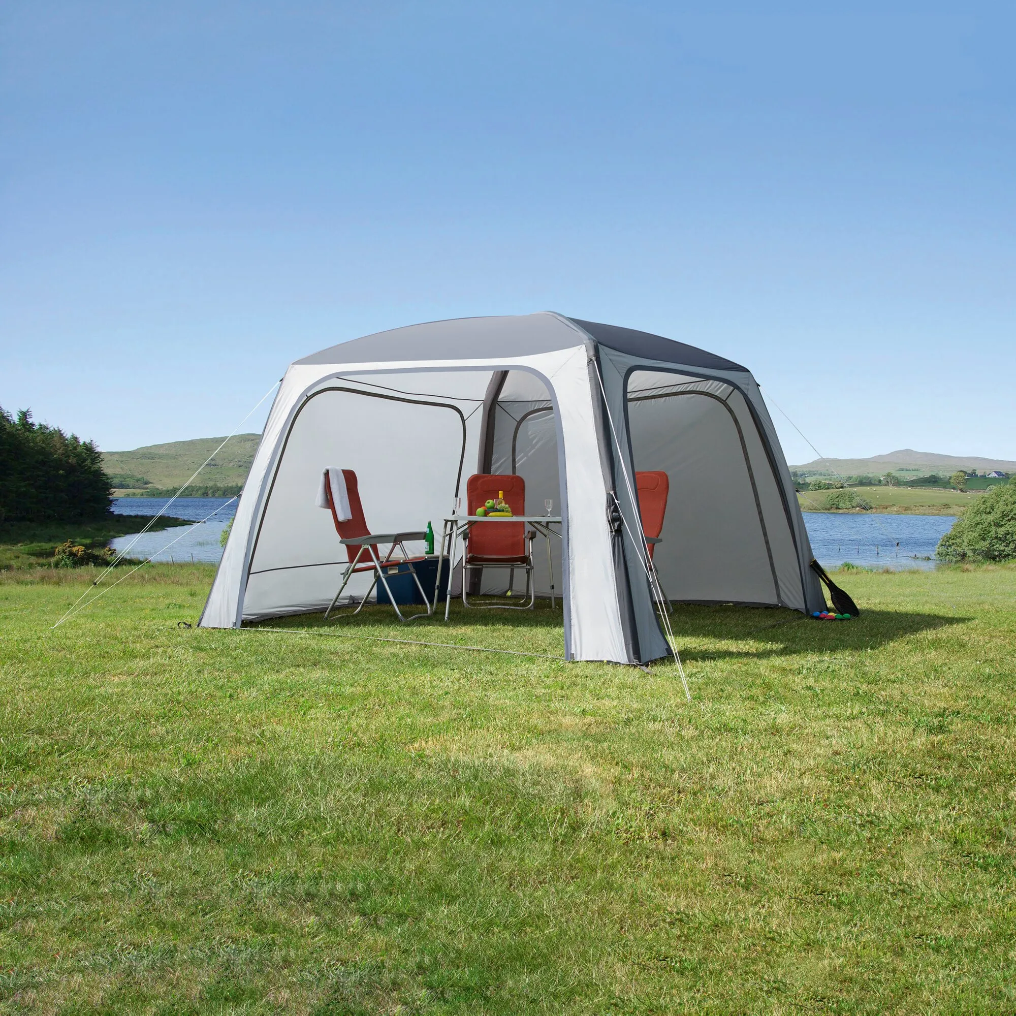Pavillon Relax Air in Grau mit geöffneten Seitenwänden, roten Campingstühlen und Tisch auf einer Wiese am See