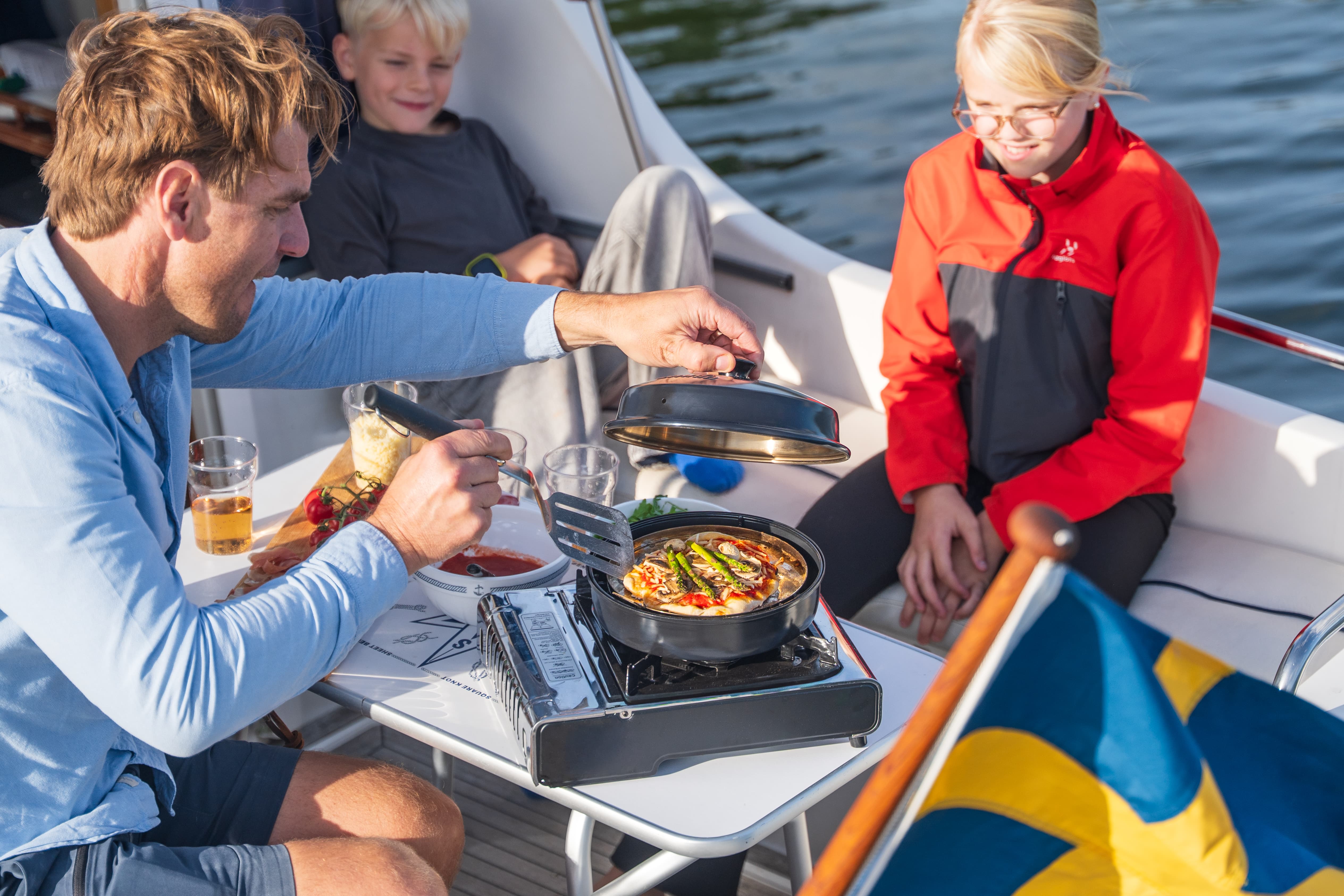 Auf einem Boot bereitet eine Gruppe Pizza zu; ein Mann in blauem Hemd öffnet den Deckel eines tragbaren Stovetop-Pizzaofens, daneben Pizza