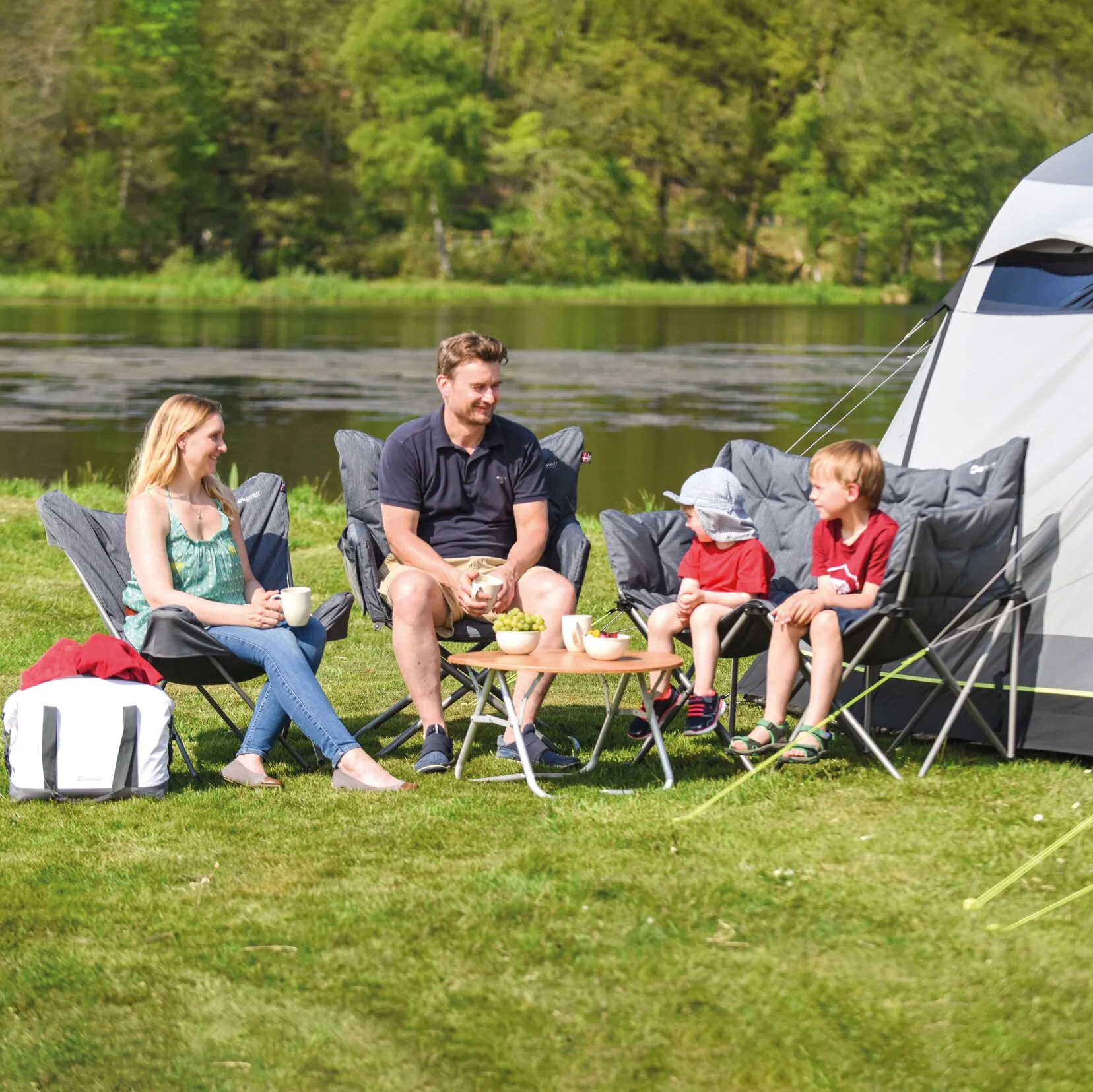 Familie mit zwei Kindern sitzt am See in grauen Mondstühlen der Möbelserie Lake vor einem Zelt und einem Campingtisch