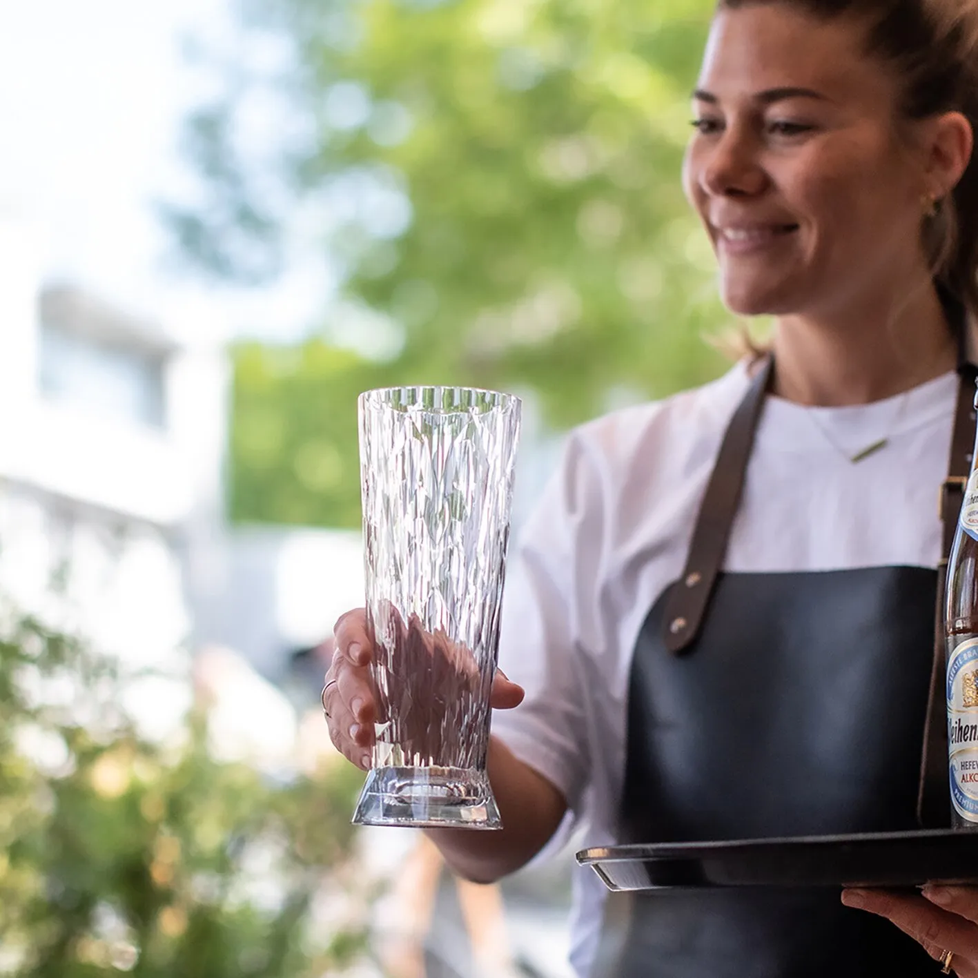 Servicekraft hält ein hohes, transparentes Weißbierglas mit facettiertem Muster und breitem Fuß auf einem Tablett gras