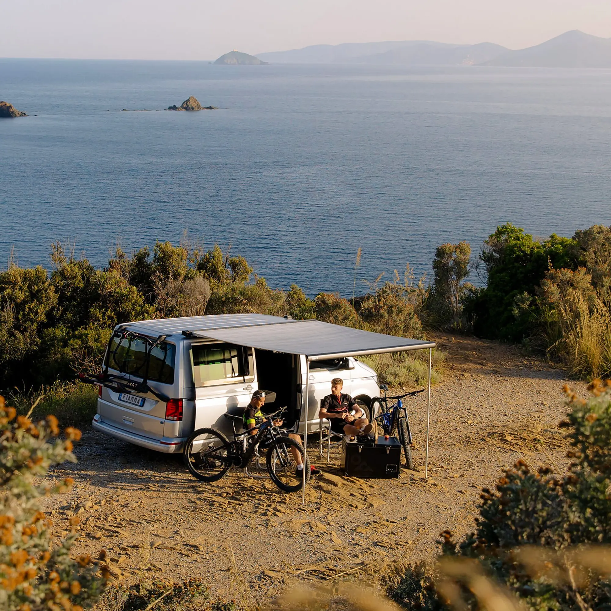 Weißer VW T6 mit dachmontiertem Carry-Bike Pro Deep Black Fahrradträger, darauf montierte Fahrräder, Markise ausgefahren, Küste