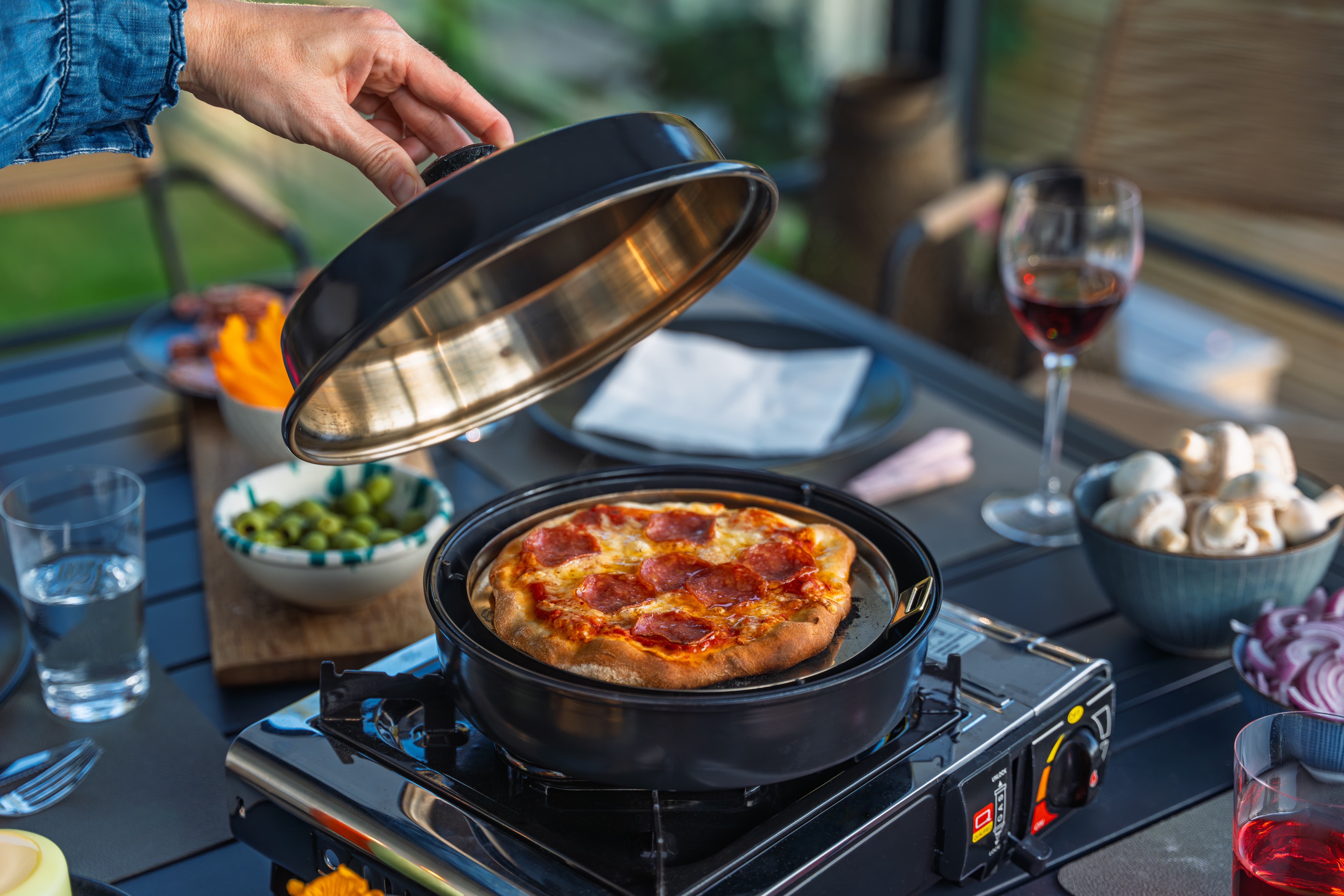 Eine Person hebt den Deckel eines schwarzen Stovetop-Pizzaofens; darunter Pizza mit Pepperoni, Oliven und Brotrollen am Tisch im Freien