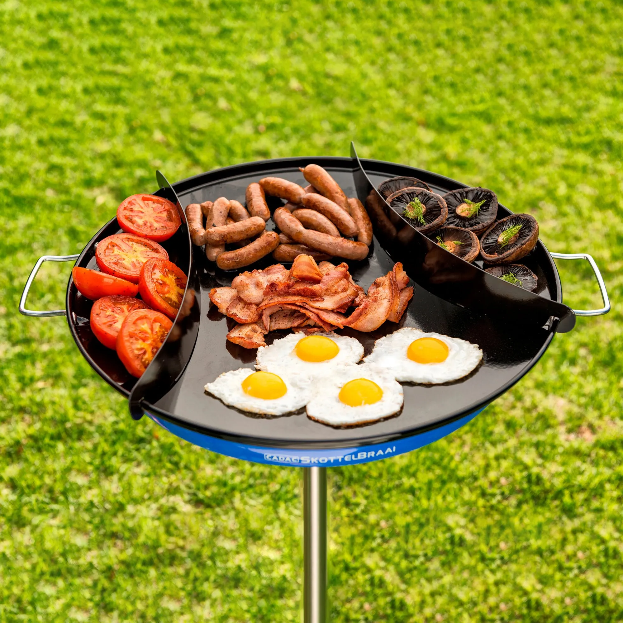 Schwarze runde Warmhalteschienen Skottel auf einem Grill mit Würstchen, Speck, Spiegeleiern, Tomaten und Pilzen belegt
