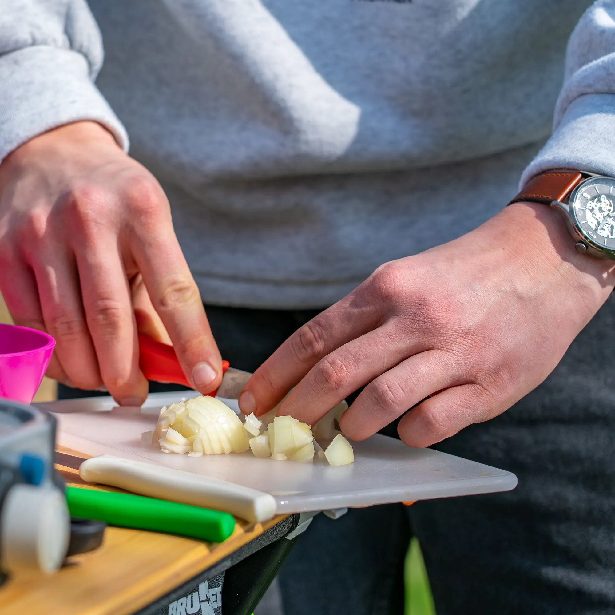 Eine Person in grauem Hoodie hackt Zwiebeln auf einem Schneidebrett aus dem Schneidebrett-Set; pinker Messbecher und Werkzeuge daneben