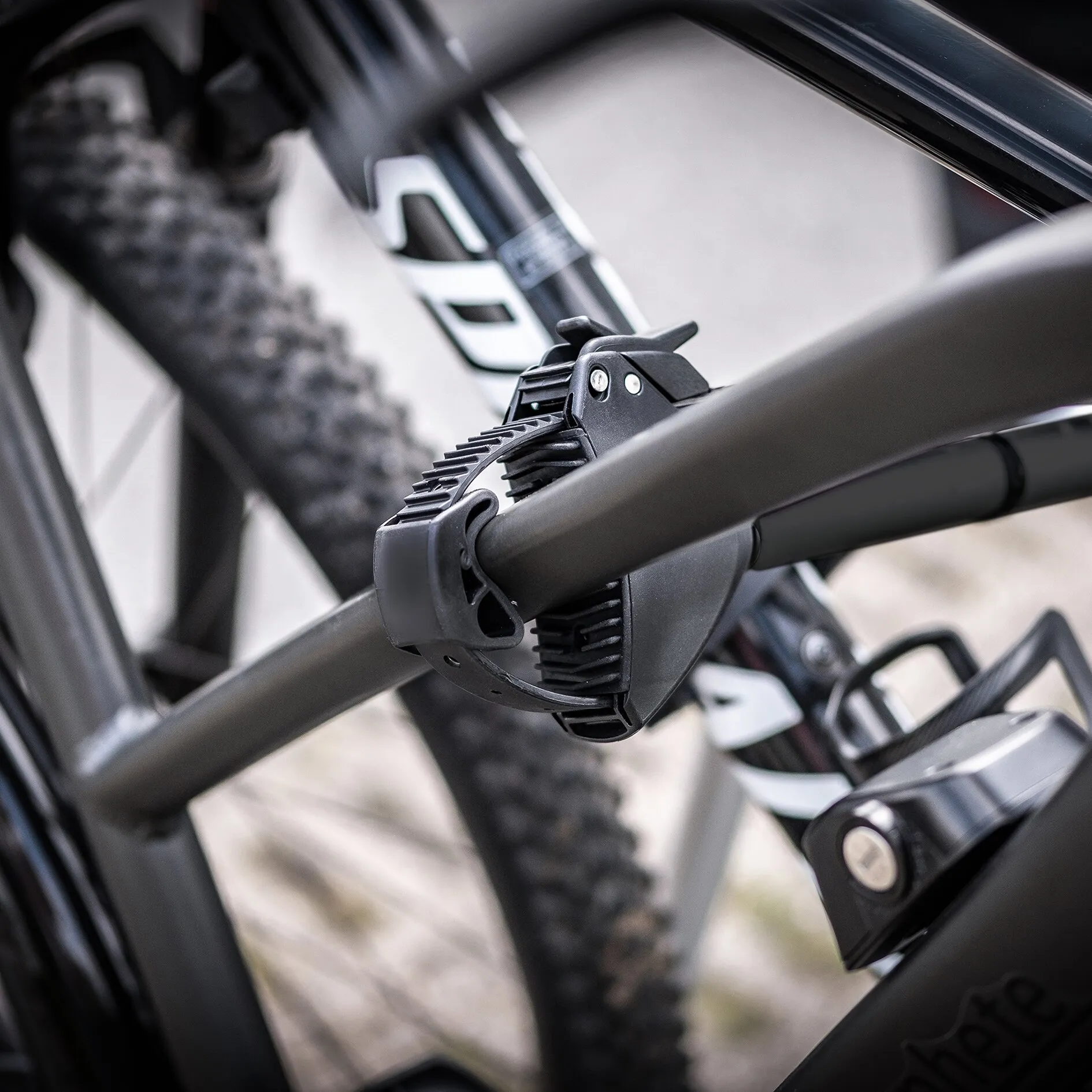 Schwarzer Fahrradträger mit Anhängerkupplung, Nahaufnahme der Klemmvorrichtung am Rahmenteil, unscharfer Mountainbike-Reifen im Hintergrund
