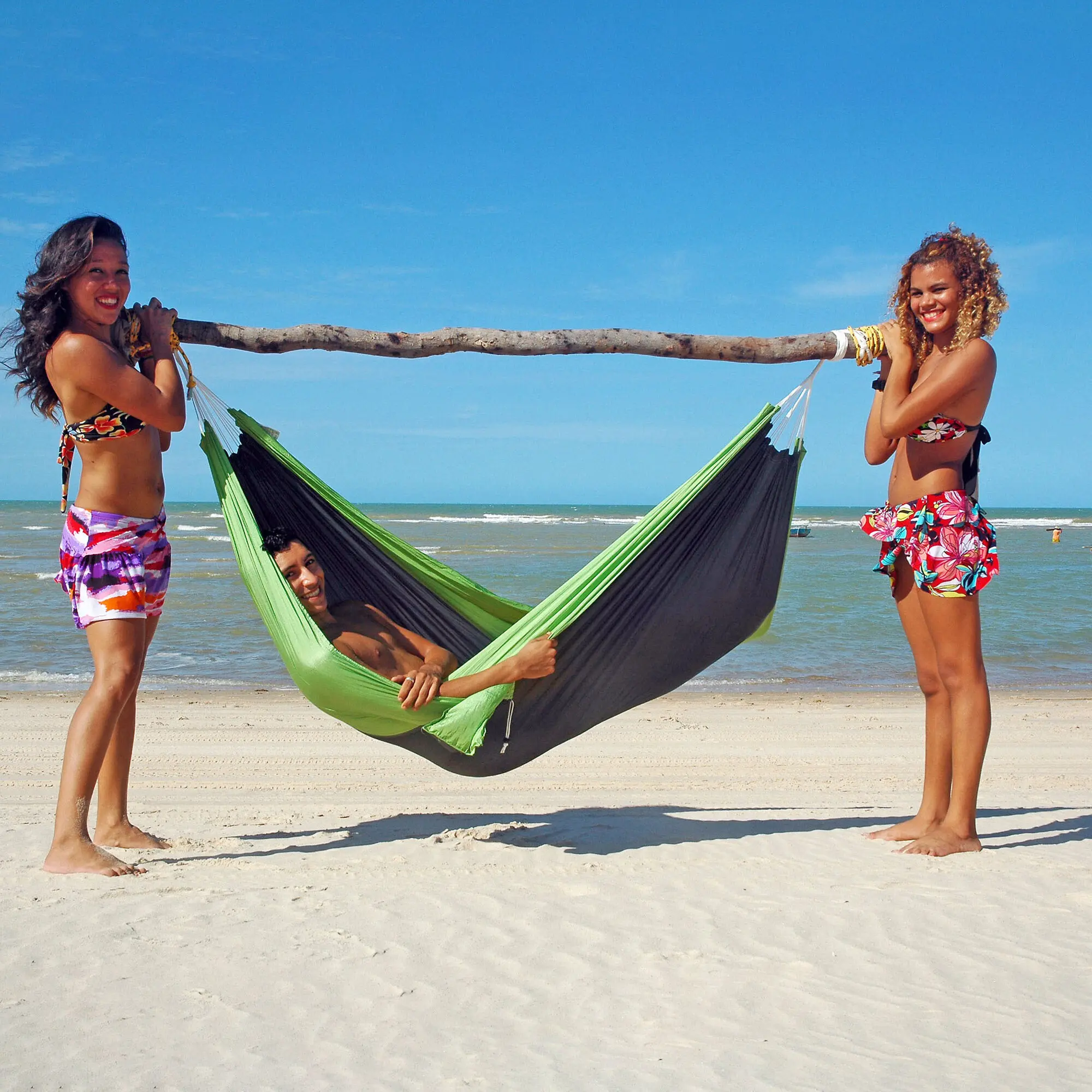 Hängematte Silk-Traveller forest grün-schwarz wird von zwei Frauen am Strand festgehalten, eine Person liegt darin