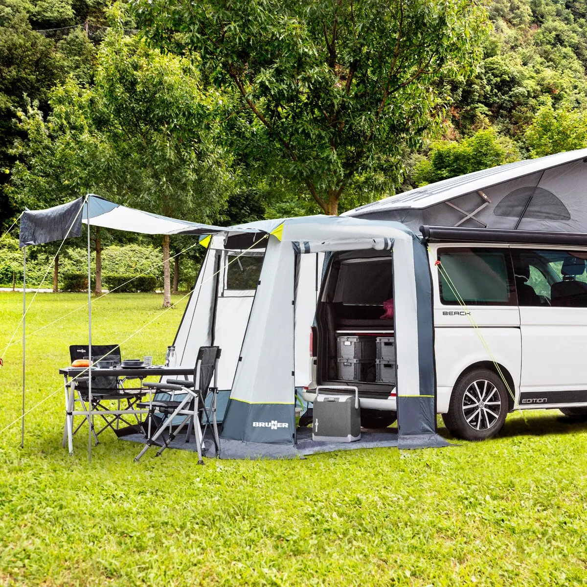Graues Brunner Heckzelt Comet am geöffneten Heck eines VW T5/T6 mit Sonnendach, Campingtisch und Stühlen auf einer Wiese