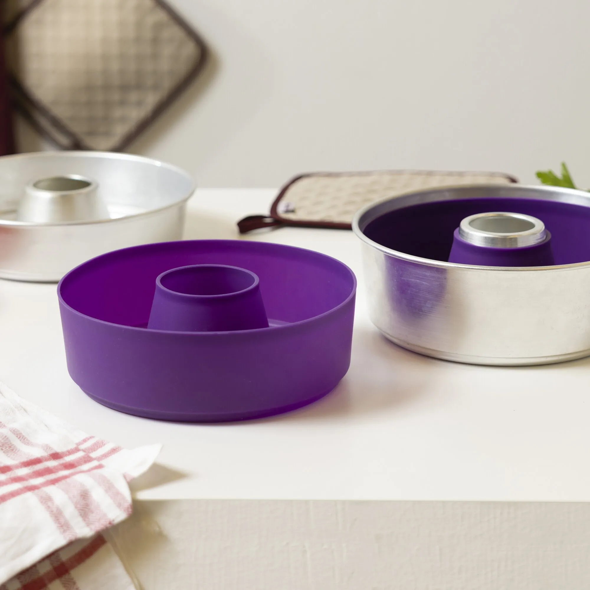 Violette runde Silikonform Maxi 3L mit Mittelrohr, neben passenden Metallbackformen auf einer Küchenarbeitsfläche