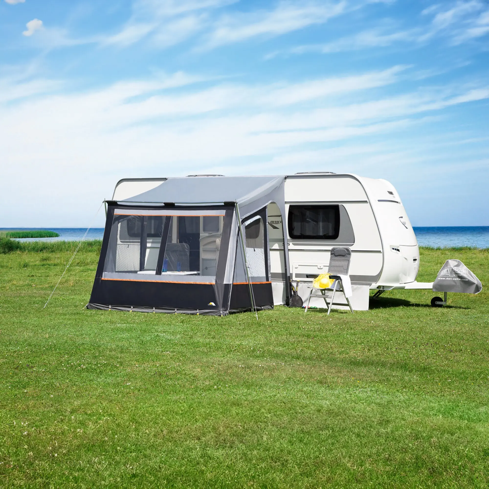 Fortuna II Teilzelt angebaut an weißen Wohnwagen, Vorderzelt mit Netzfenstern und orangefarbener Rand, Gras, Meer und blauer Himmel im Hintergrund
