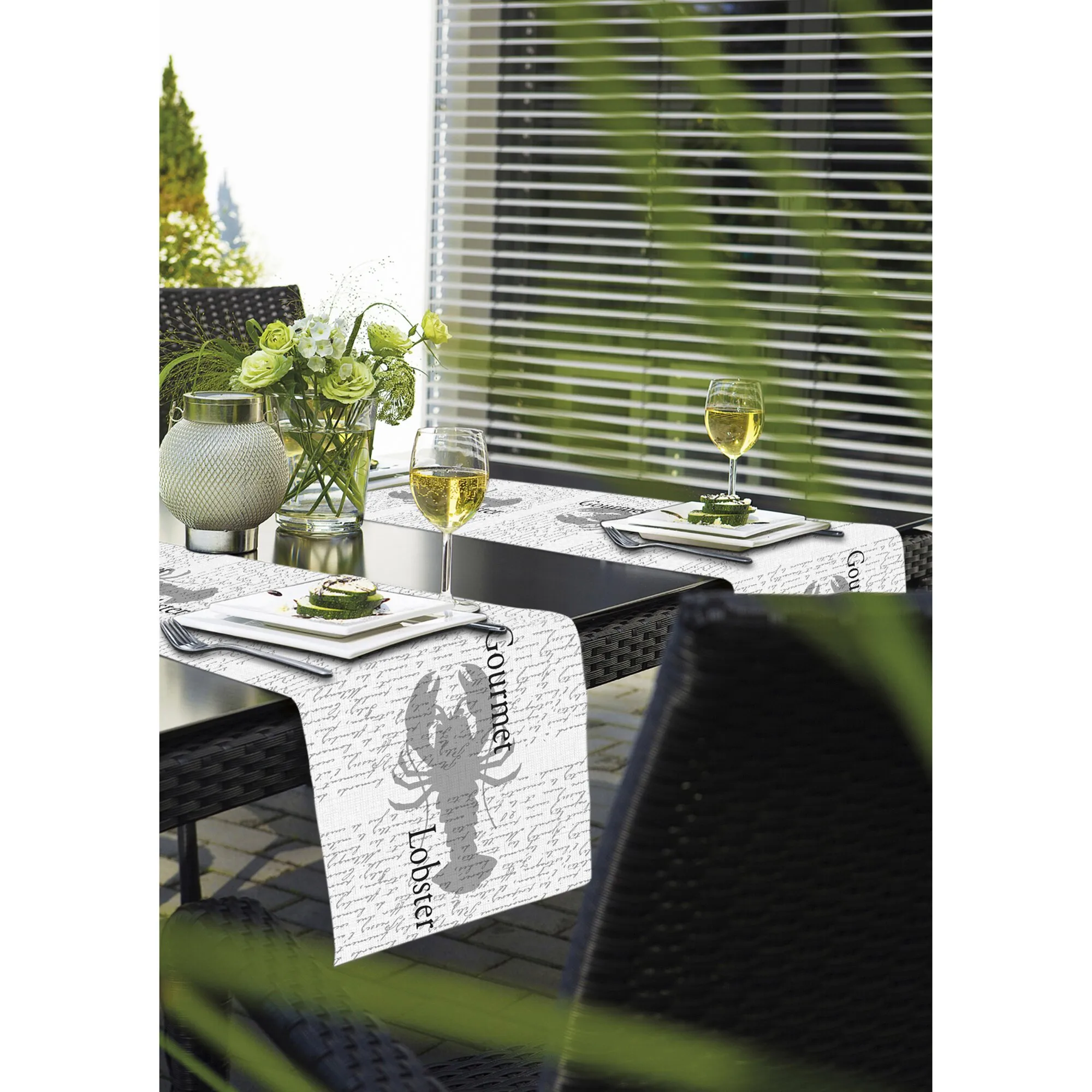 Weißer Tischläufer Miami mit grauem Schrift- und Motivaufdruck auf einem gedeckten Gartentisch mit Blumen und Weingläsern