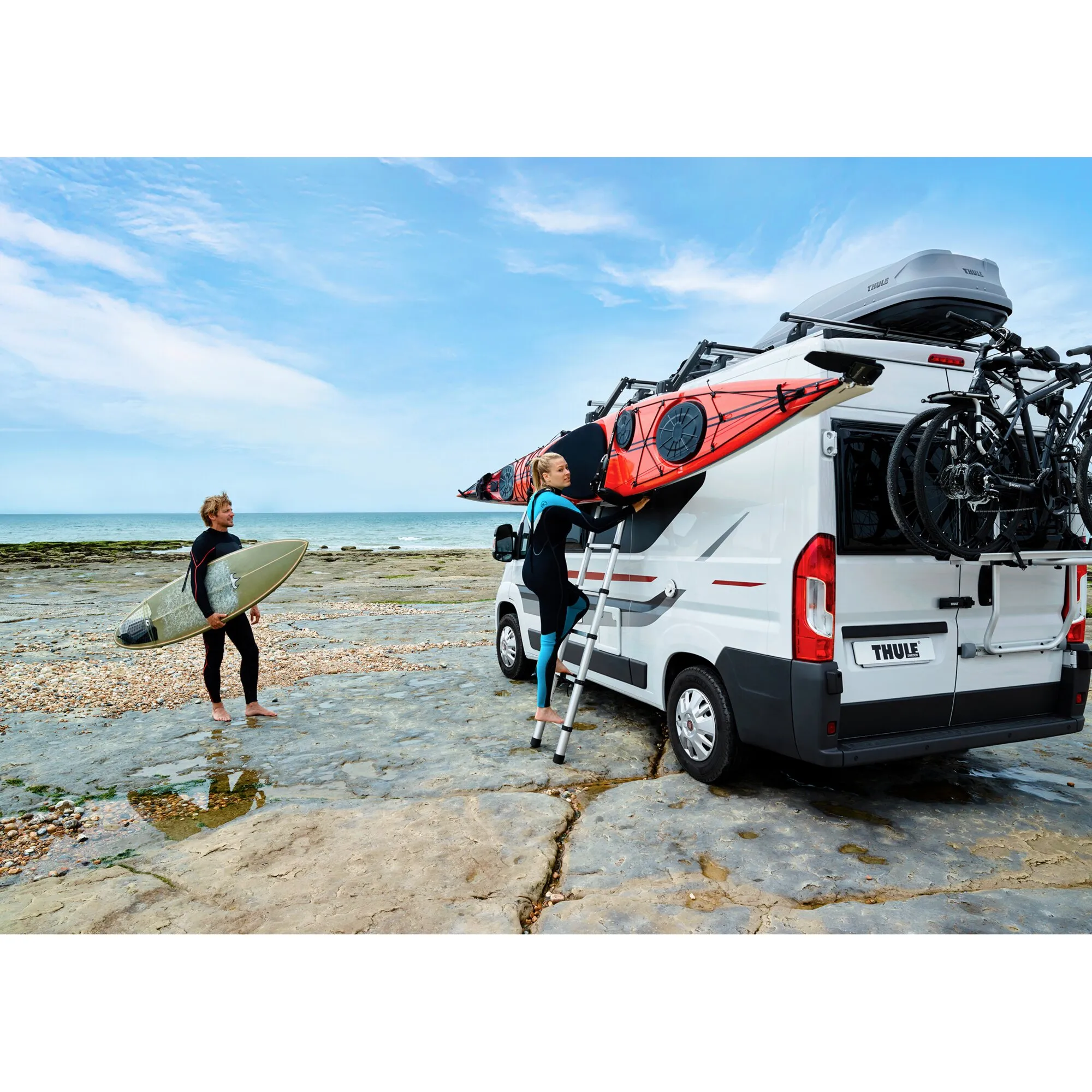 Thule Hullavator Pro am Wohnmobil: Eine Frau lädt ein rotes Kajak auf den Dachträger, hinten sind Fahrräder befestigt, am Strand