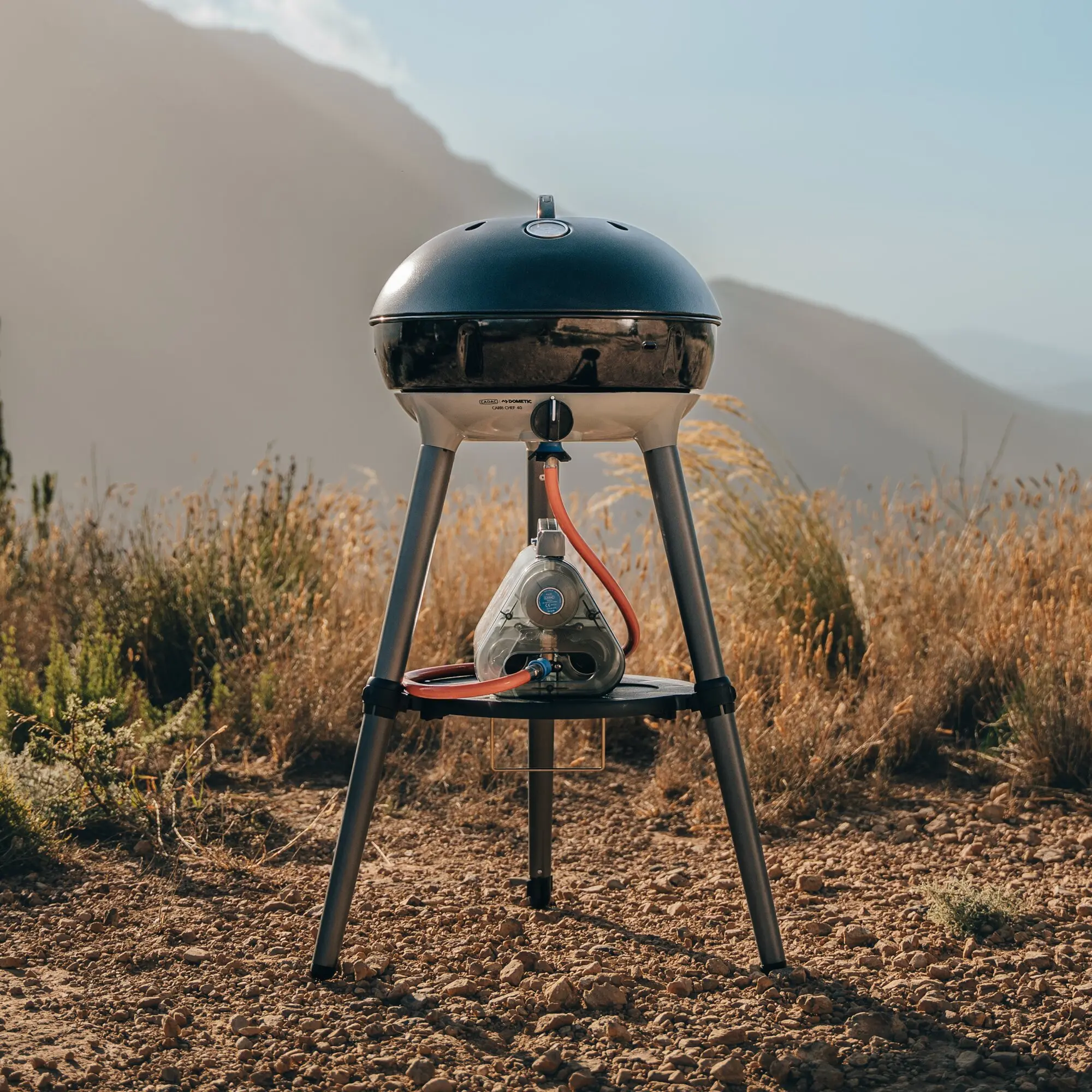 Gasgrill Carri Chef 40 mit dunkelblauem, halbkugelförmigen Deckel auf Dreibein-Stativ im Freien roter Schlauch führt zur Gaszufuhr unten