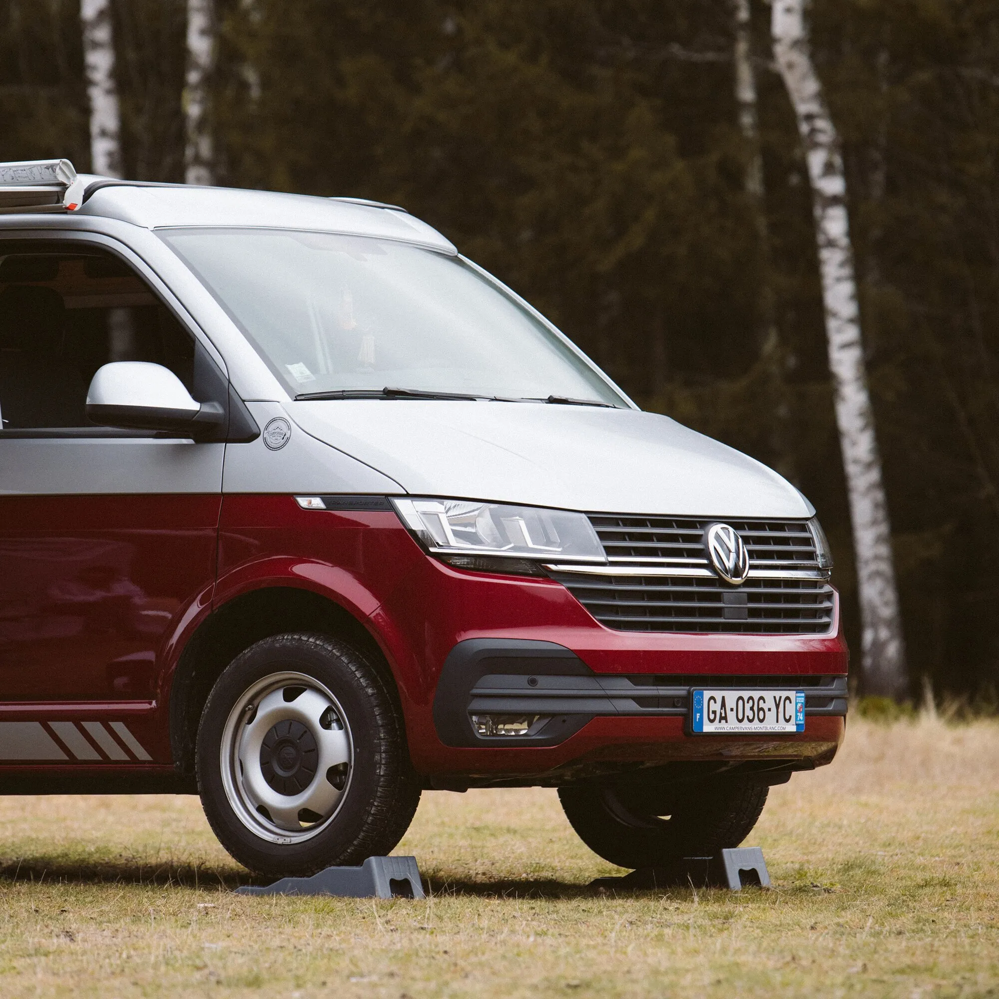Rot-weißer Camper steht mit den Vorderrädern auf zwei grauen Auffahrkeilen Level Up auf einer Wiese vor einem Wald