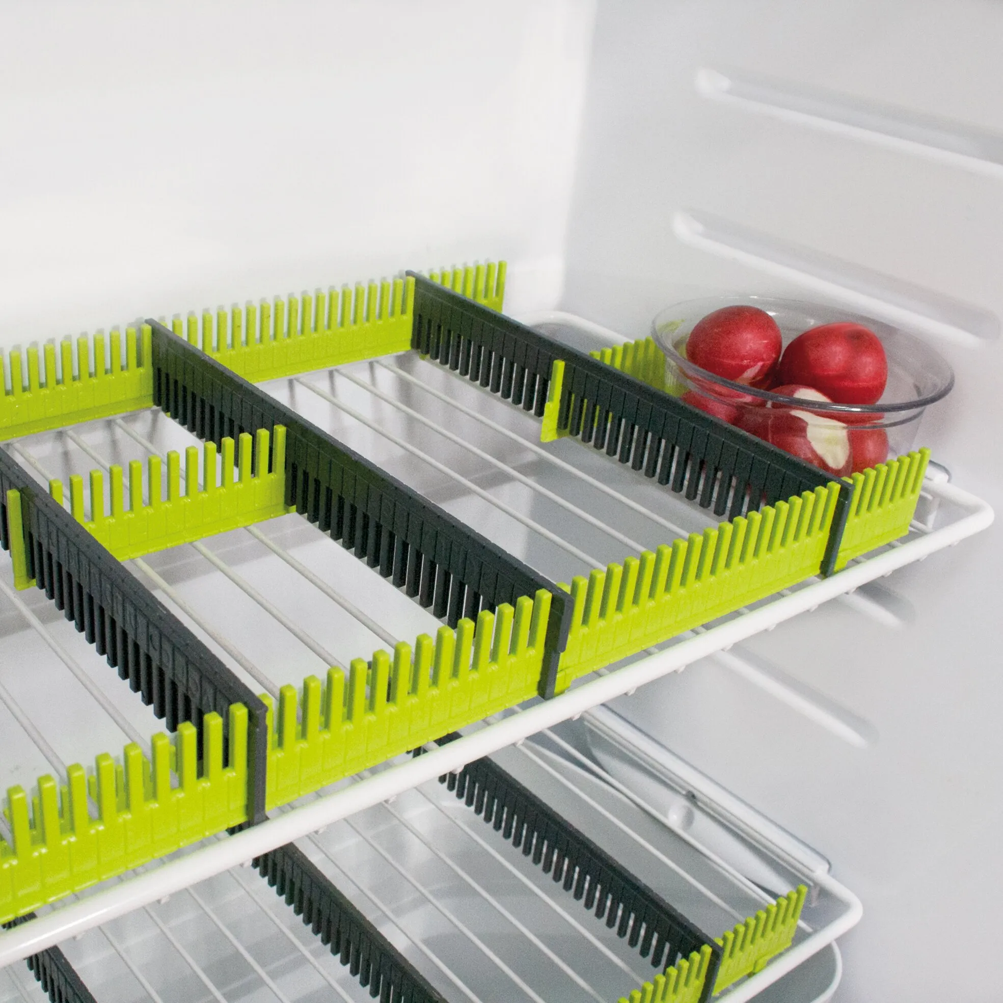Limegrüne und schwarze Stauleisten auf Kühlschrankgittern, mit einer Glasschale roter Äpfel im Hintergrund
