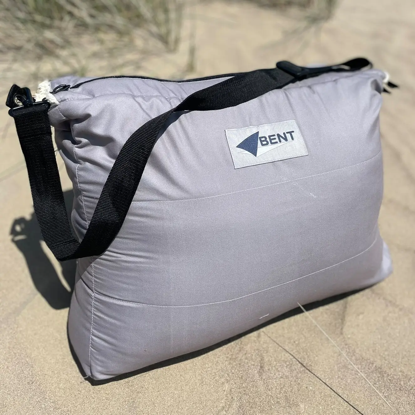 Grauer Vanlife Schlafsack Bentcho in leichter Hülle mit schwarzem Tragriemen liegt auf Sand, Logo-Aufnäher Bent sichtbar