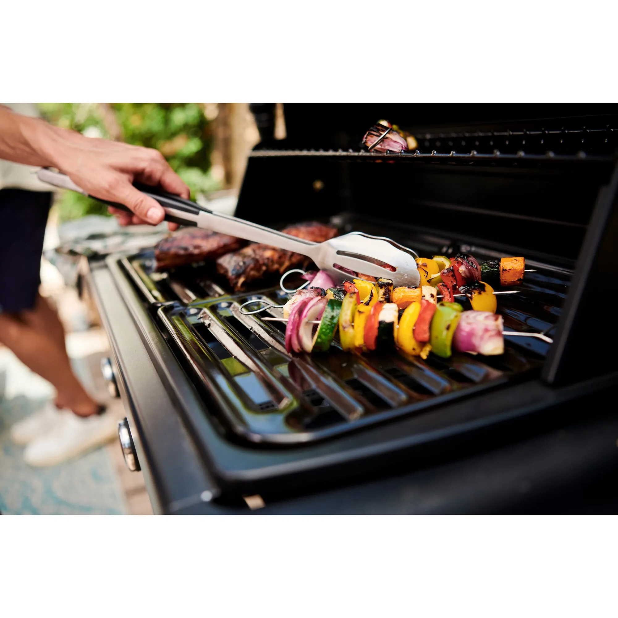 Geöffneter schwarzer Gasgrill Compact 3 L mit Fleisch und bunten Gemüsespießen auf dem Rost, eine Hand nutzt eine Grillzange