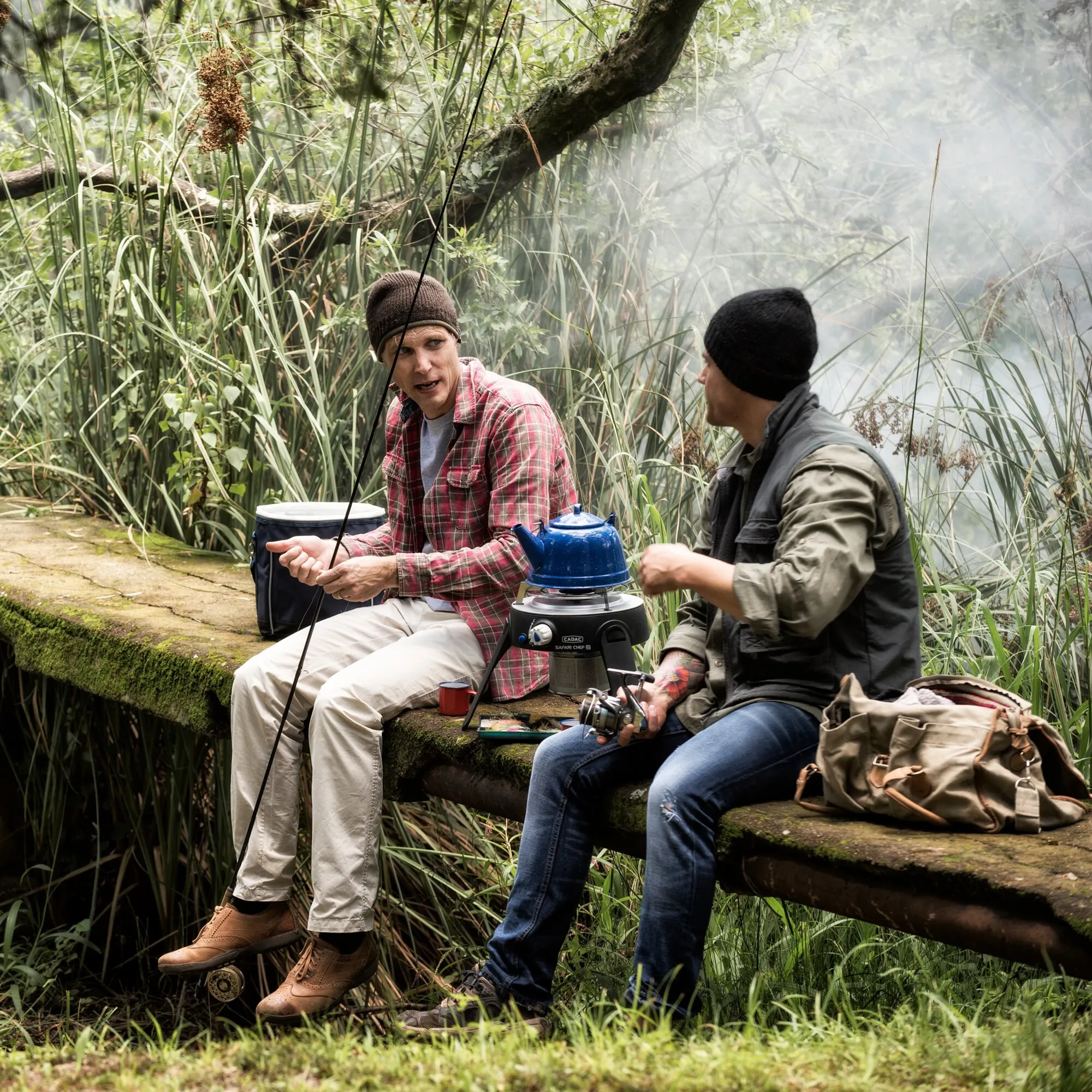 Zwei Angler sitzen im Schilf auf einem Baumstamm; zwischen ihnen steht ein schwarzer Gasgrill Safari Chef 30 mit blauem Wasserkessel