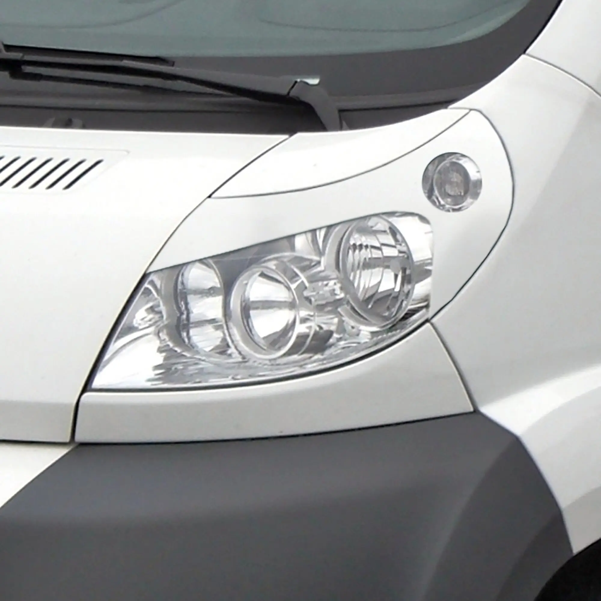 Weiße, glänzende Scheinwerferblende Böser Blick installiert über dem Scheinwerfer im Frontbereich eines Fiat Ducato, Windschutzscheibe mit Wischer sichtbar