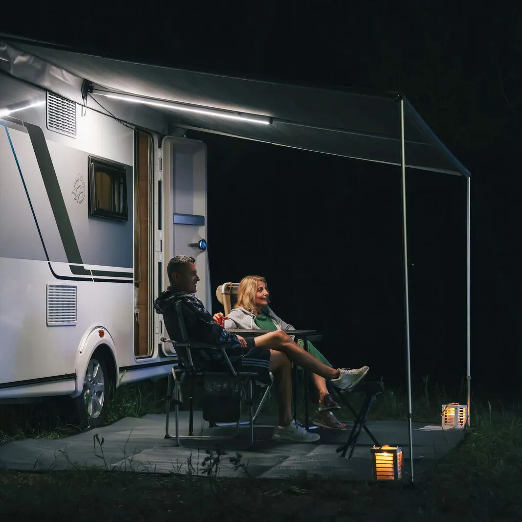 Wohnwagen mit ausgefahrener Markise und integrierter LED-Spannstange, darunter zwei Personen beim Camping am Abend