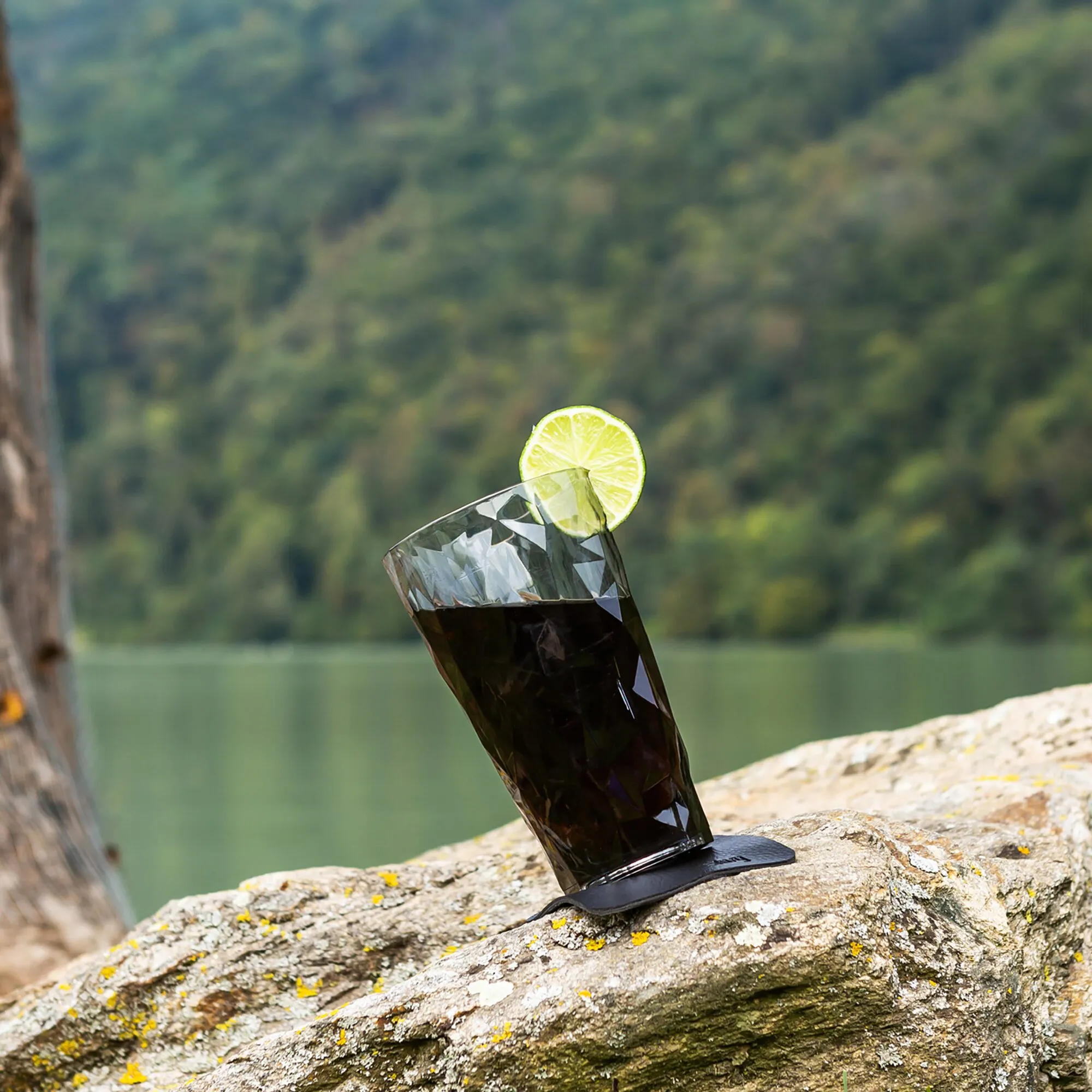 Graues, facettiertes Longdrinkglas aus Kunststoff mit dunklem Getränk, Limettenscheibe und magnetischem Untersetzer auf einem Felsen