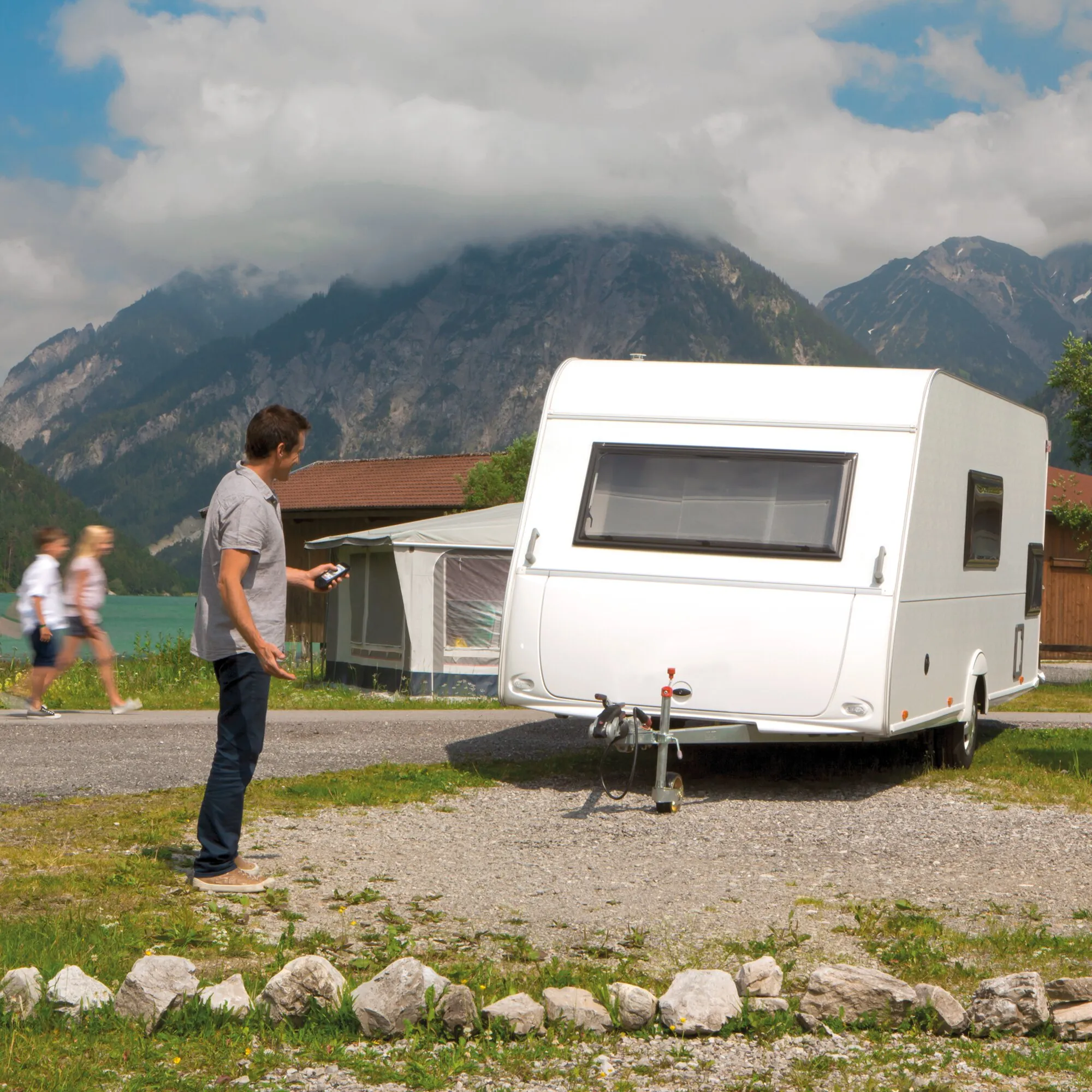 Mann steuert einen weißen Wohnwagen mit Rangierantrieb per Handgerät auf einem Campingplatz vor alpiner Bergkulisse