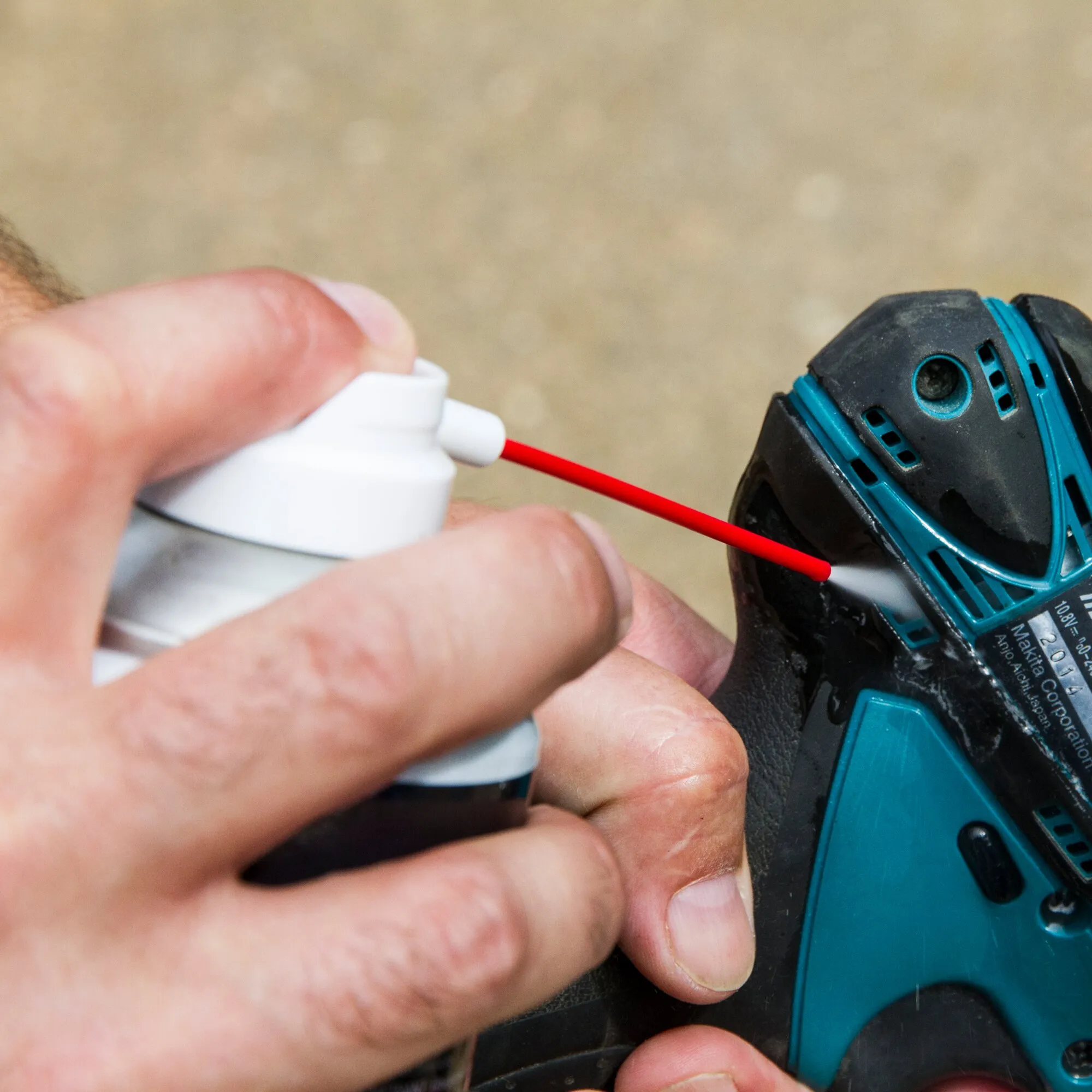 Hand sprüht Multifunktionsspray GT7 mit rotem Sprührohr auf ein blau-schwarzes Akkuschrauber-Gehäuse
