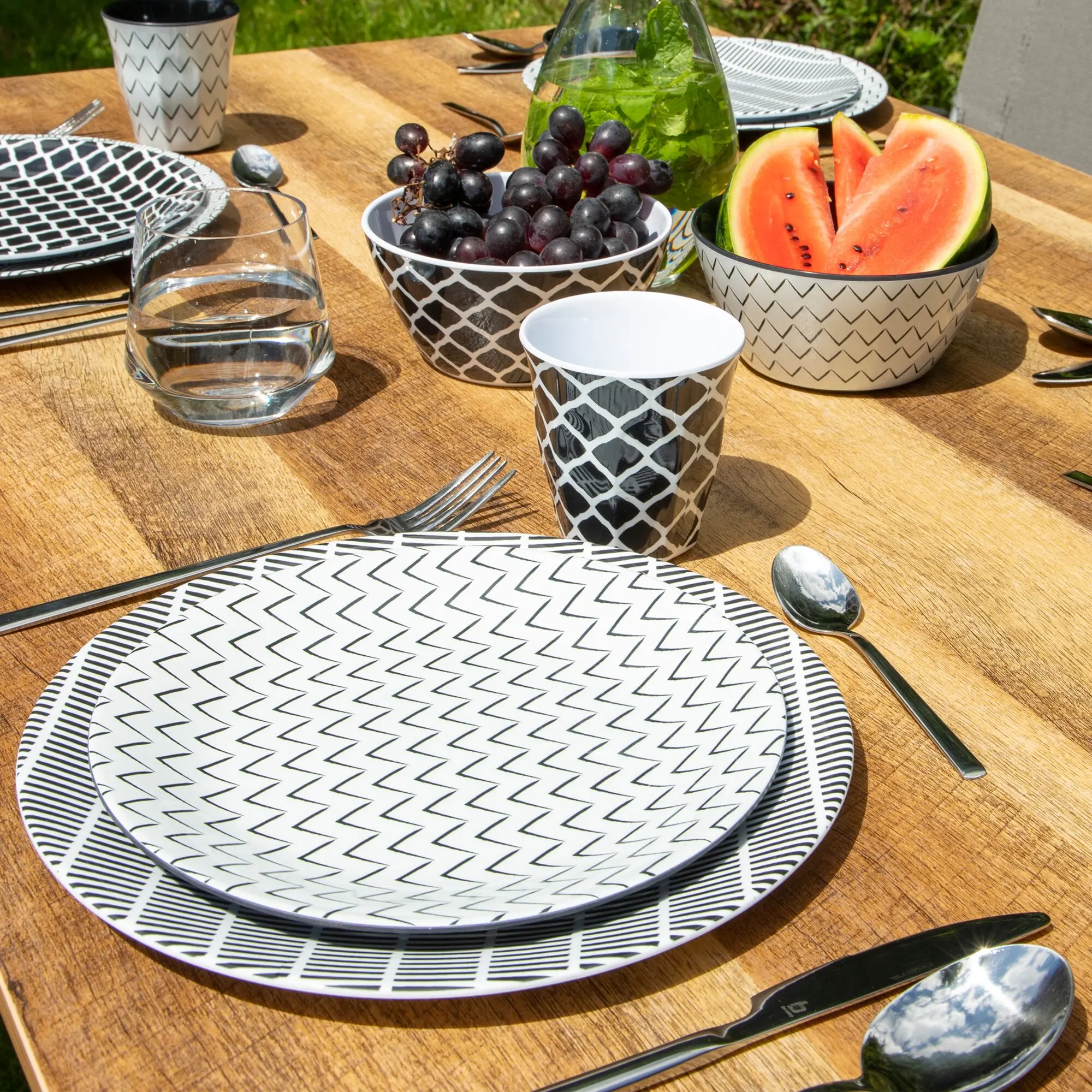 Schwarz-weiße Mix & Match 16-teilig Geschirrserie mit Zickzack-Muster, Teller, Schale und Becher auf Holztisch, Obstschüssel mit Trauben und Wasserkaraffe daneben