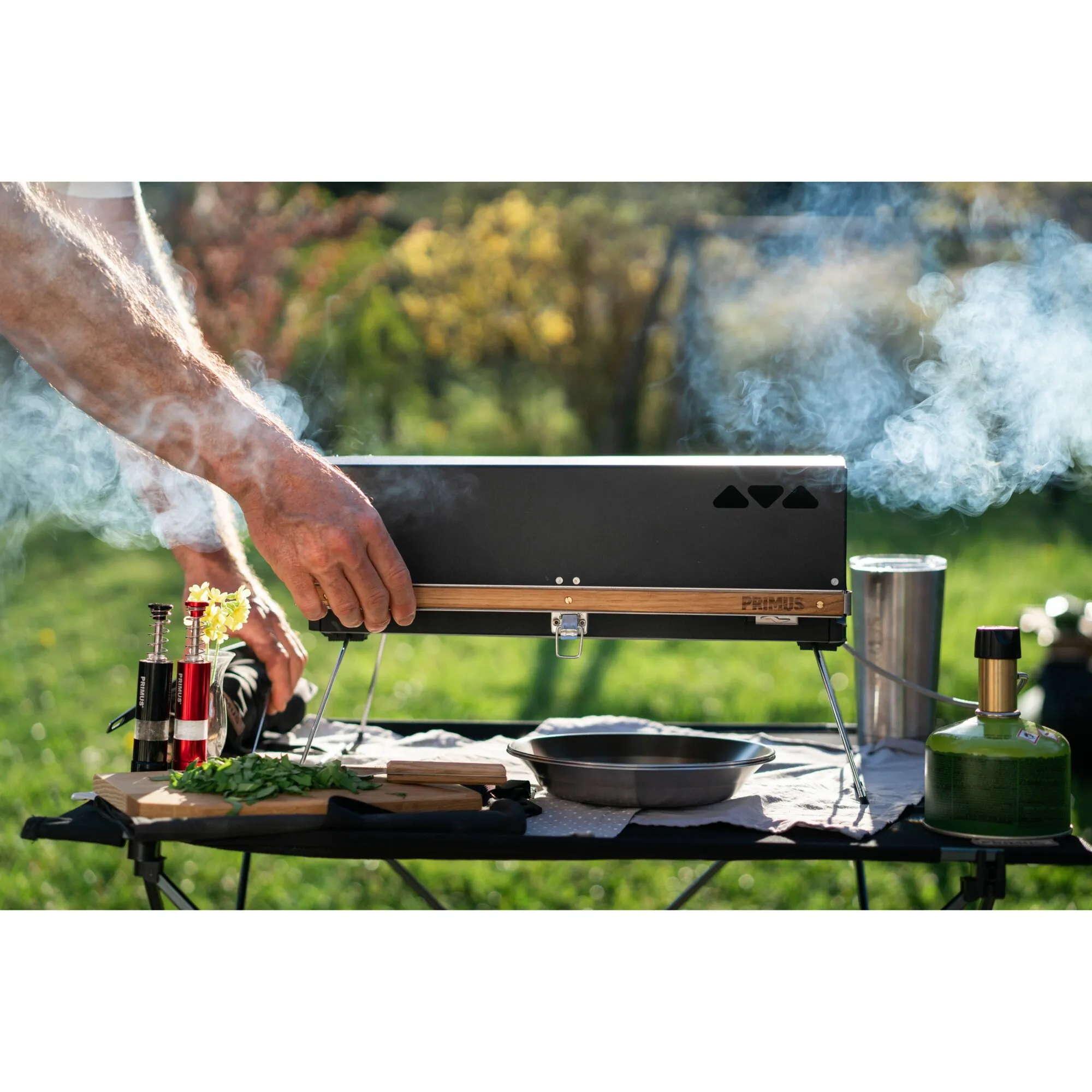 Tragbarer schwarzer Primus Kuchoma Gasgrill mit Holzleiste beim Grillen im Freien, umgeben von Rauch, Kochutensilien und Gasflasche