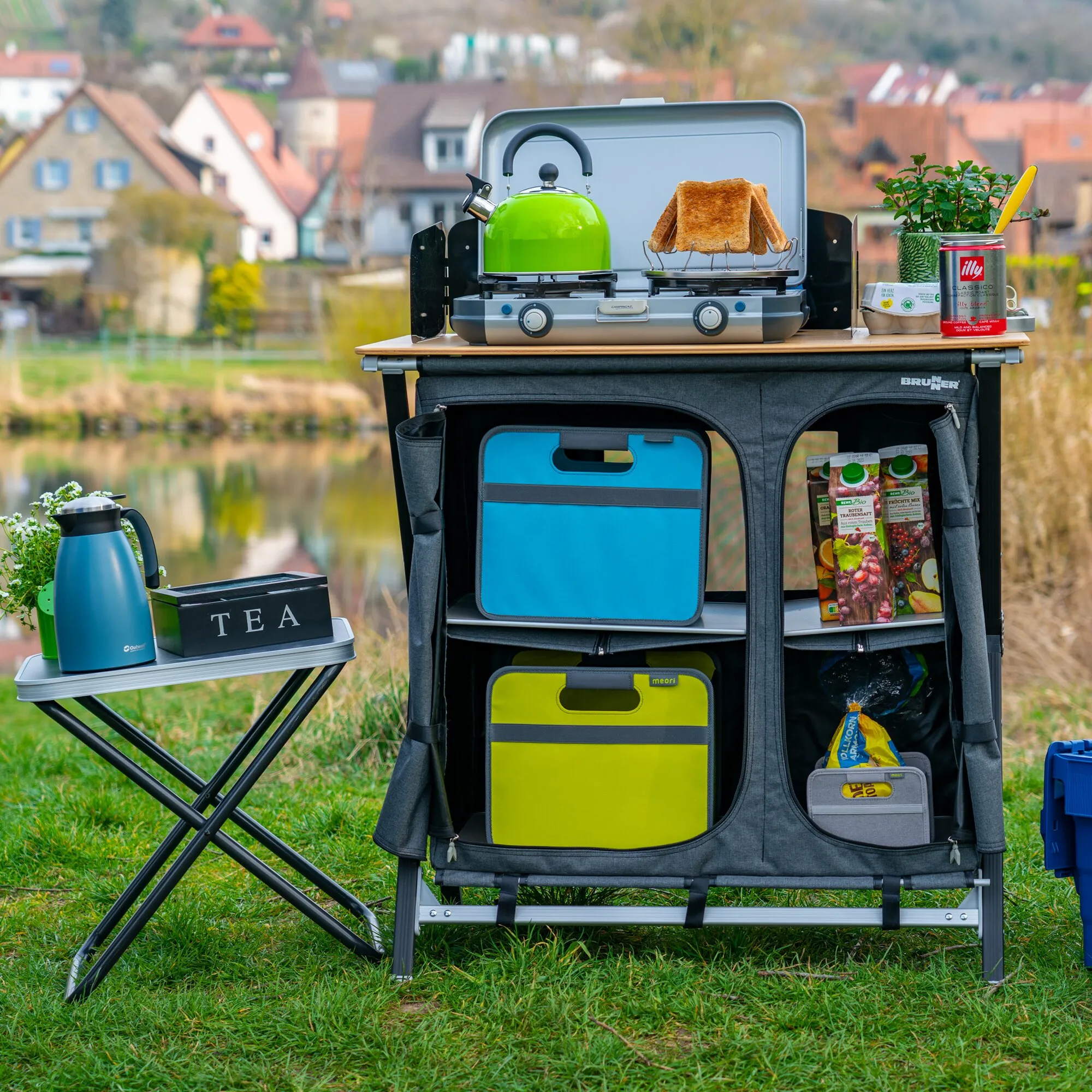 Grauer Vielzweckschrank Mercury Cross Square als Campingküche mit Ablage, Fächern und tragbarem Kocher auf einer Wiese