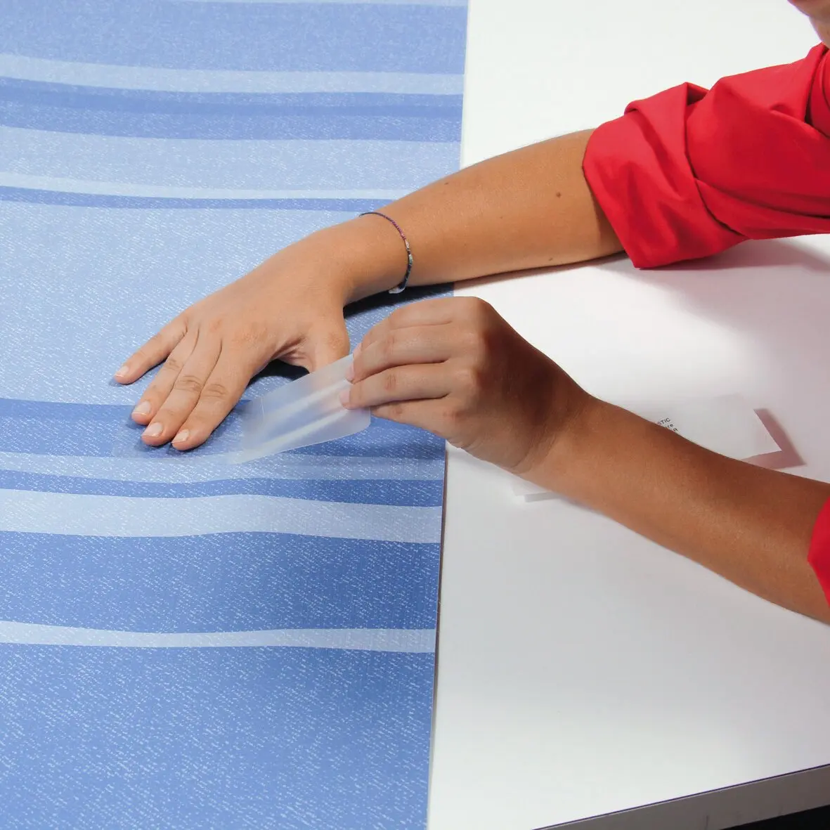 Person mit rotem Ärmel zieht eine transparente Klebefolie von einem Blatt ab und legt sie auf eine blau gestreifte Unterlage