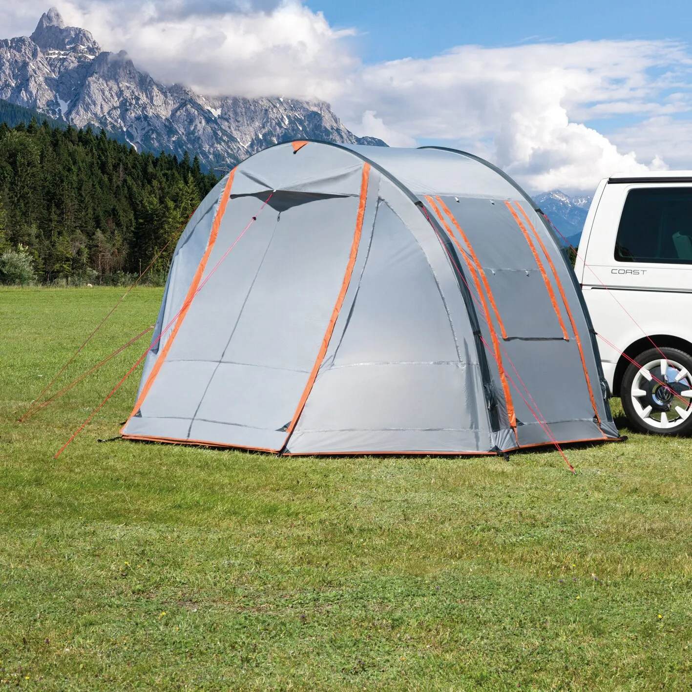 Graues Busvorzelt Picco Air mit orangefarbenen Details, aufgebaut neben einem weißen Camper auf einer Wiese vor Bergkulisse