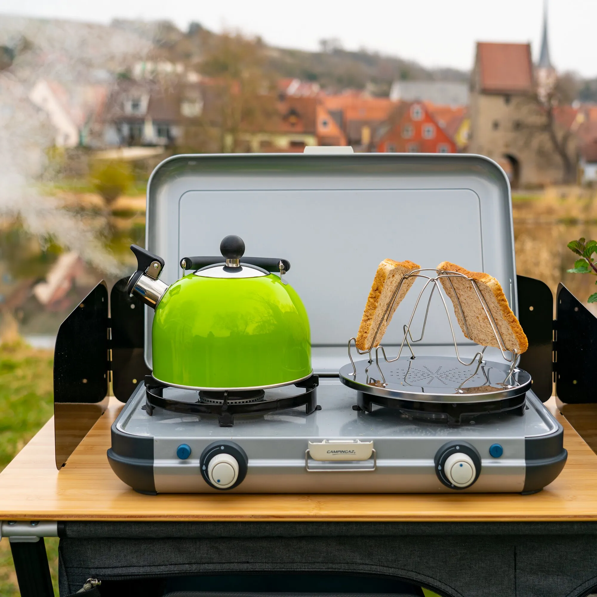 Grüner Flötenkessel Trend Color auf einem Campingkocher, daneben Toast auf einem Grillrost vor einer Stadtkulisse