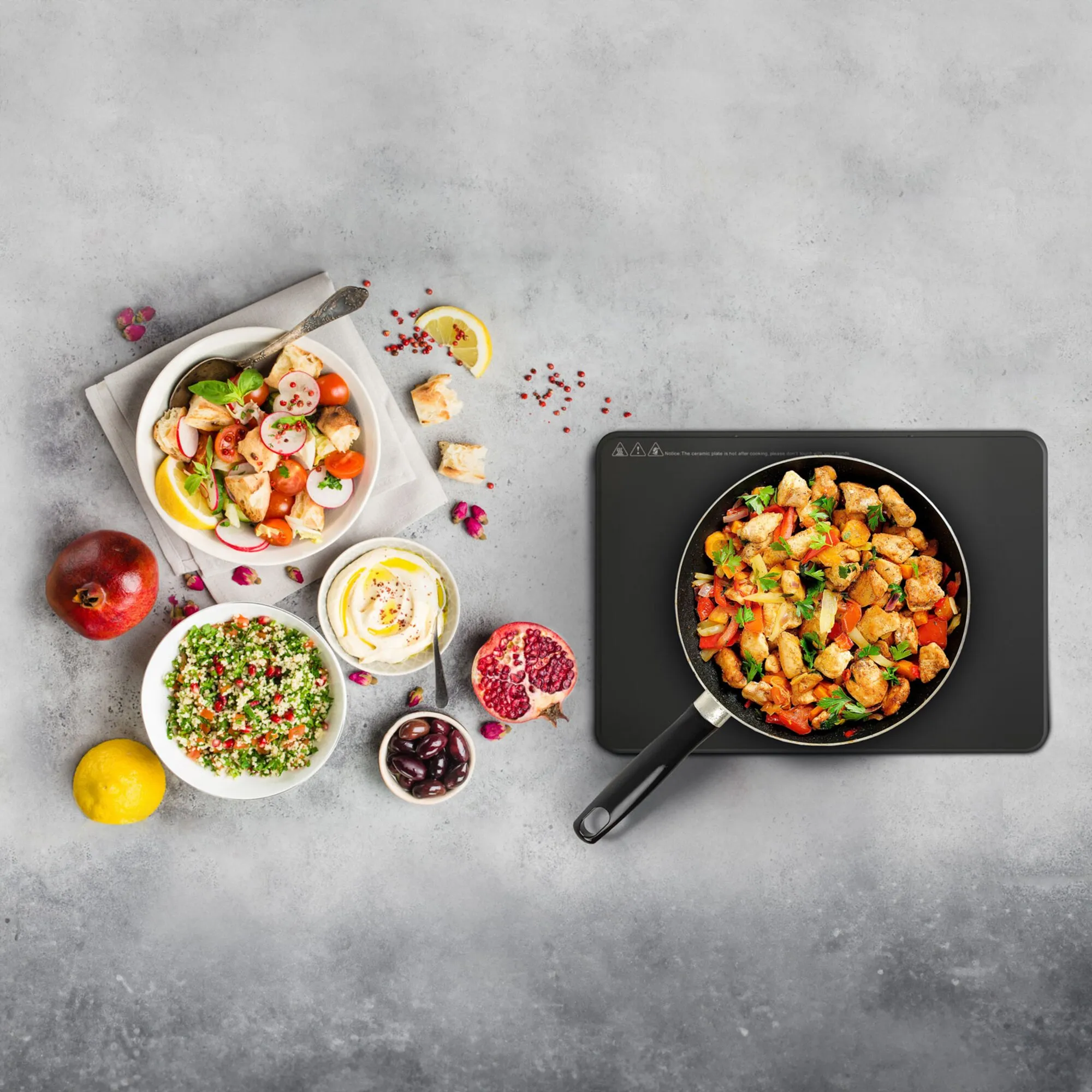 Schwarzer Induktionsgrill Hot Point mit Pfanne, in der gebratenes Hähnchen und Gemüse zubereitet werden, daneben Salate und Zutaten