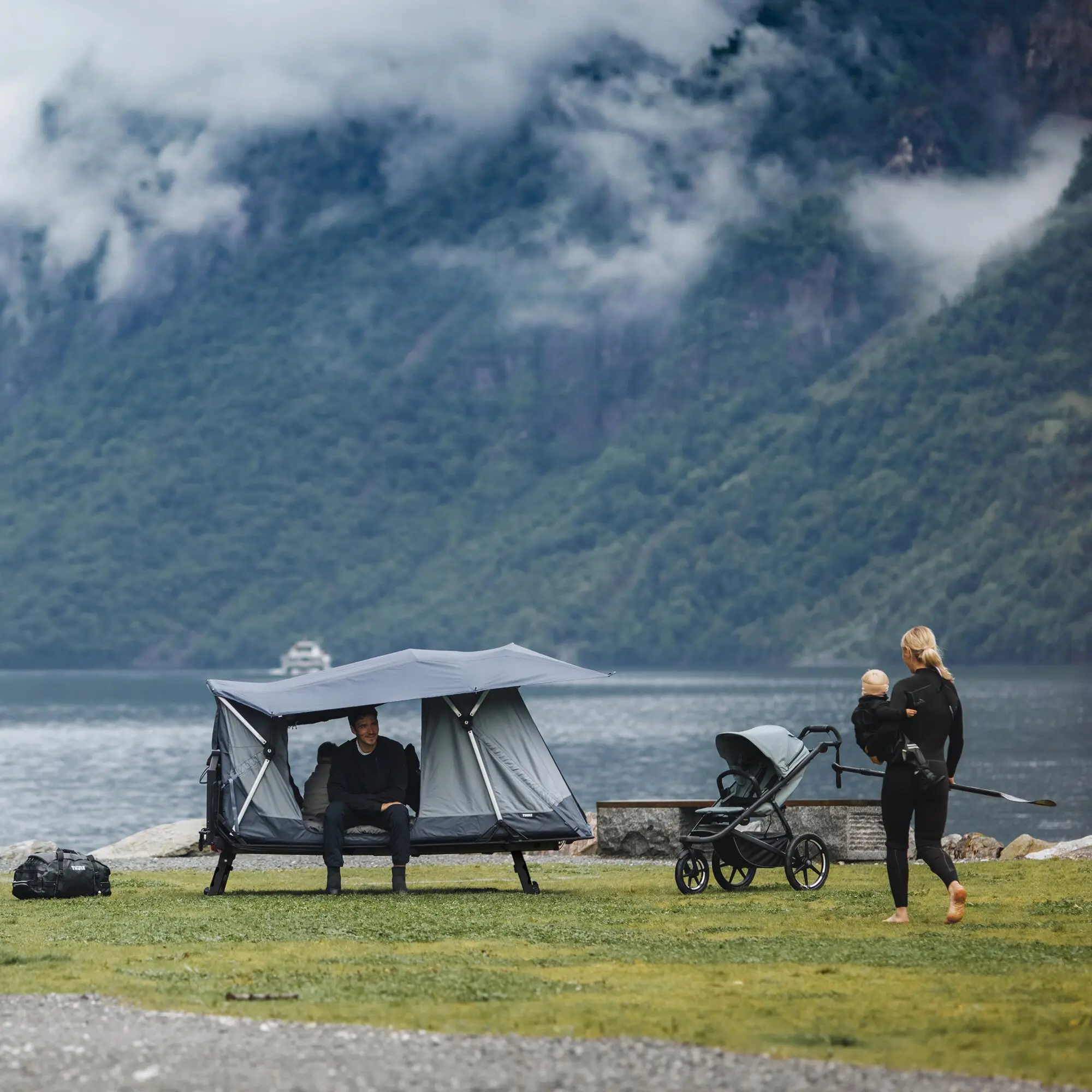 Familie am See neben einem aufgeklappten Thule Outset Anhängerkupplungszelt, graues Zelt auf Gestell, Berglandschaft im Hintergrund