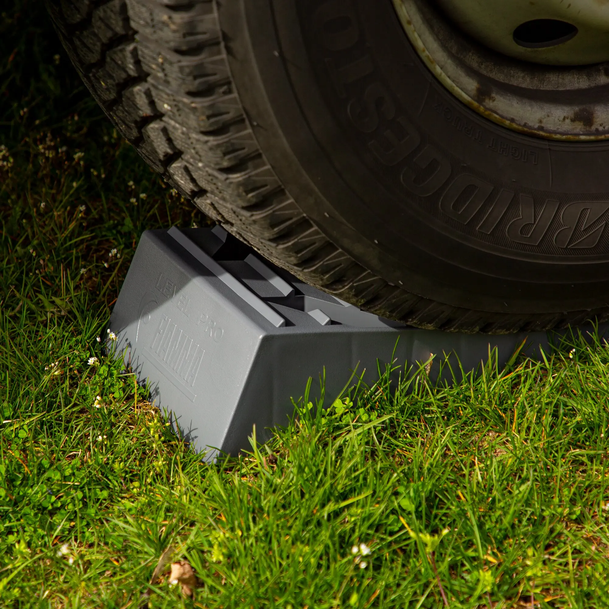 Zwei graue Auffahrkeile Level Pro 2er-Set liegen unter einem Autoreifen auf Gras, Sicht auf Profilreifen und rostige Felge