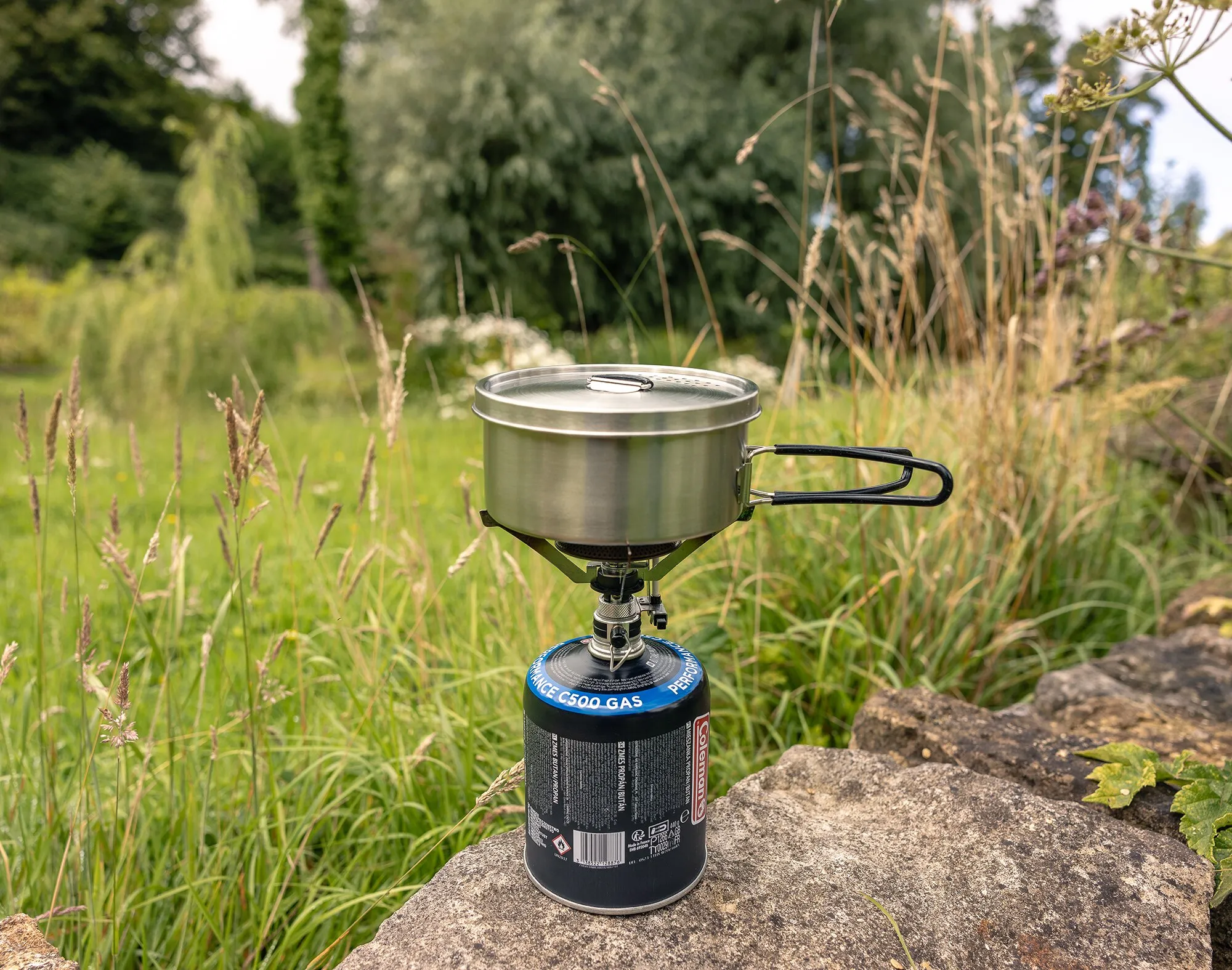 Edelstahltopf mit Deckel und klappbarem Griff auf einem Gaskocher im Trekking-Kochgeschirrset inmitten einer Wiese