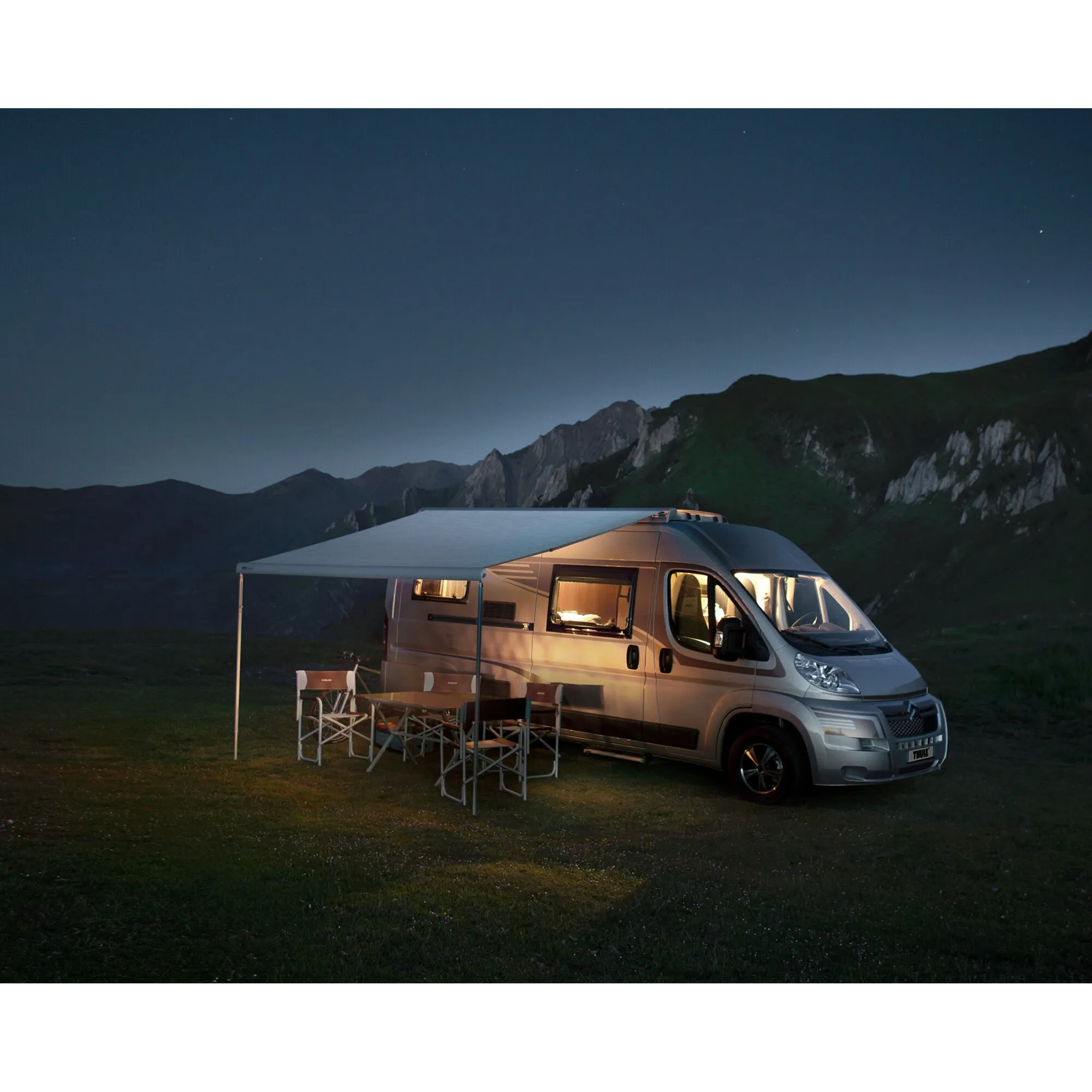 Grauer Fiat Ducato mit ausgefahrener grauer Thule Omnistor 6300 Markise und Sitzgruppe vor einer Bergkulisse bei Abendlicht
