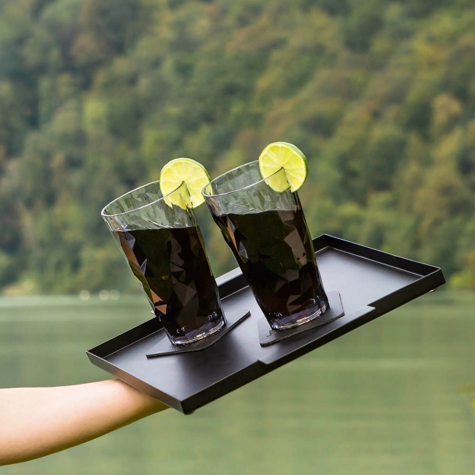 Zwei graue Longdrink-Kunststoffgläser mit dunklen Getränken und Limettenscheiben auf einem schwarzen Tablett im Außenbereich