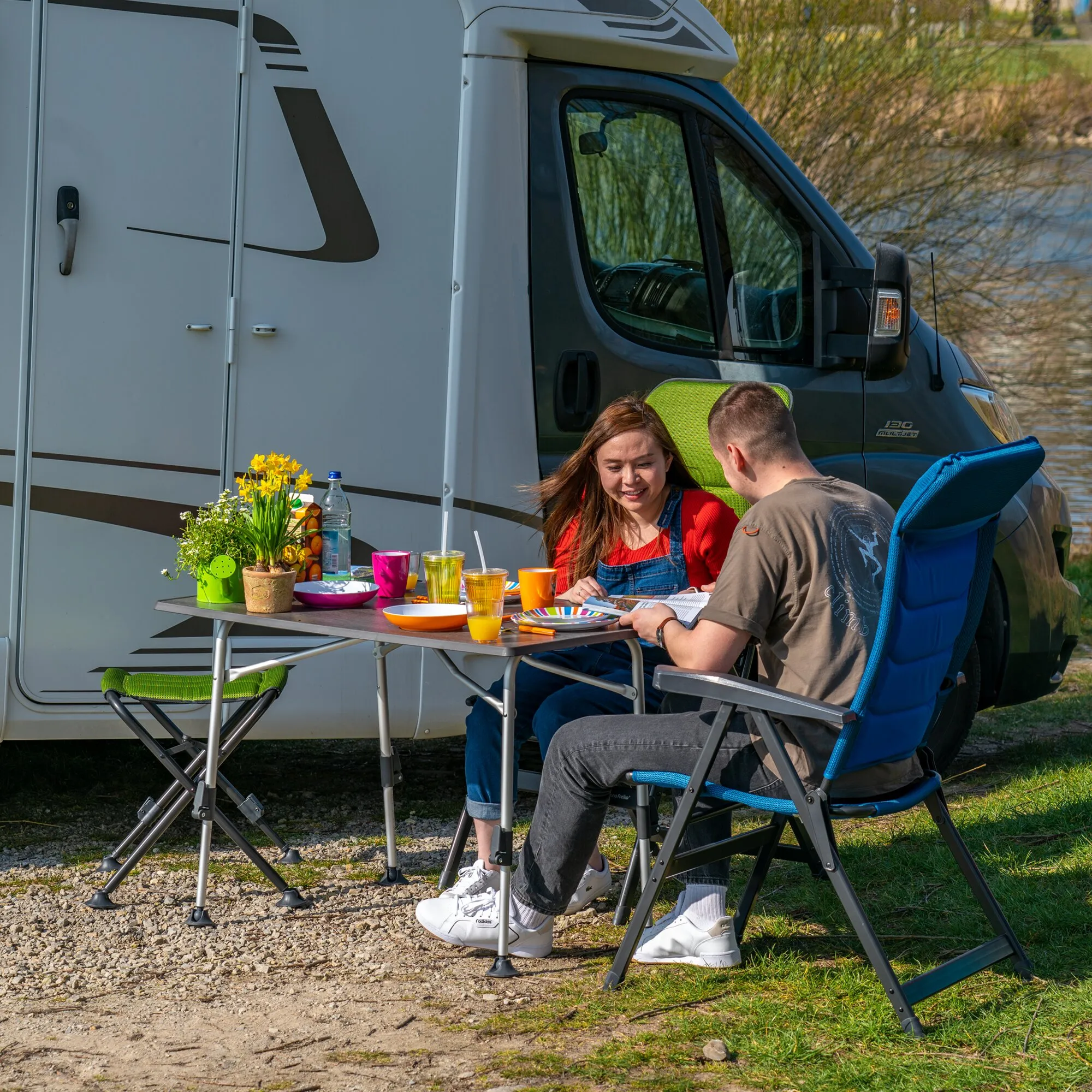 Campingtisch HighQ Blackline 115 cm mit dunkler Tischplatte, gedeckt mit Geschirr und Getränken vor einem Wohnmobil